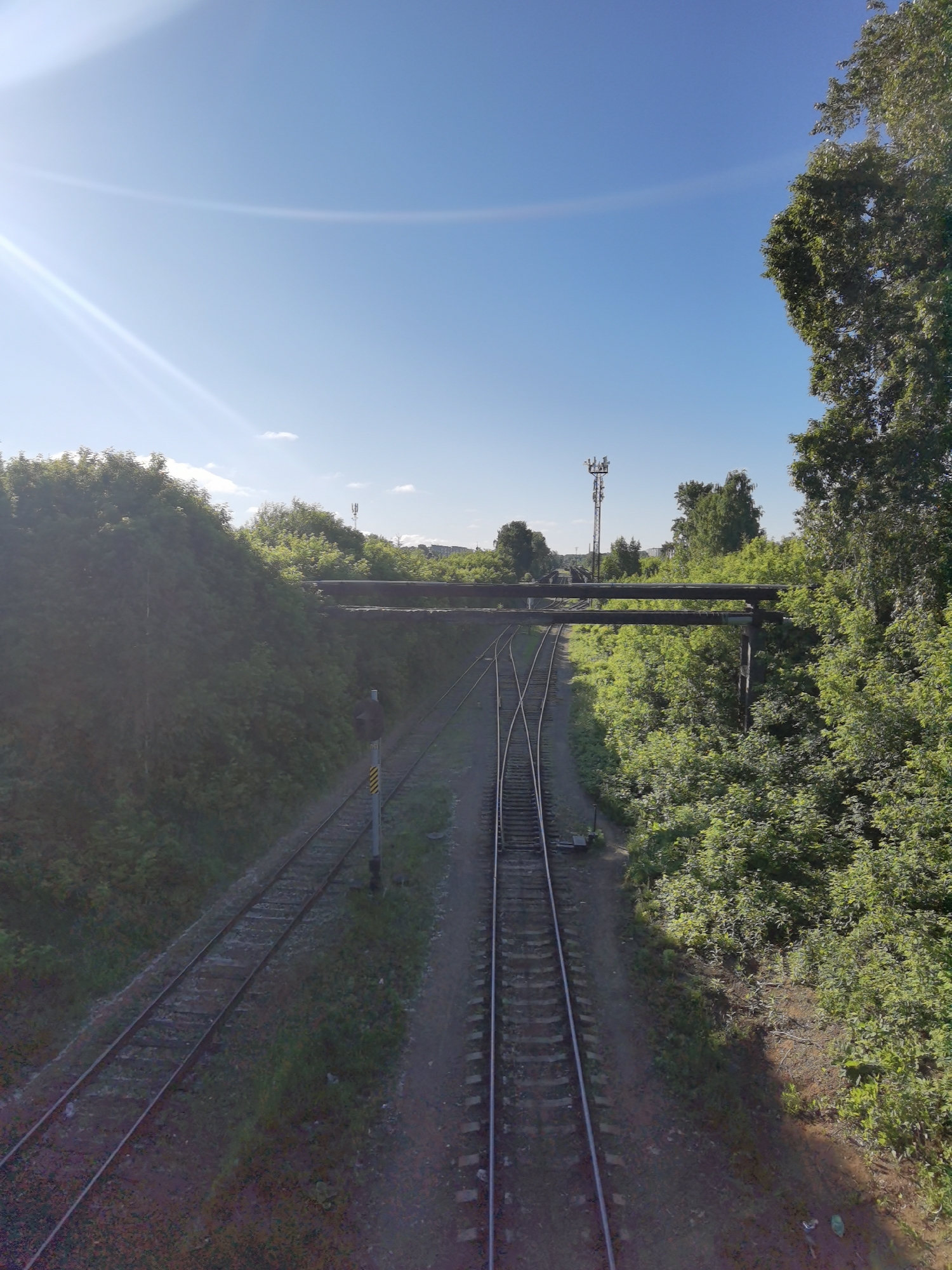 This is a ground-level, wide-angle, outdoor photograph taken on a sunny day in Chaikovsky, Russia. The scene depicts a railway track junction surrounded by lush green trees and vegetation. A signal post with yellow and black markings is visible next to the tracks. Two parallel railway tracks run into the foreground, with a third track diverging to the right, forming a junction. In the background, two horizontal pipes or conduits are suspended above the tracks, supported by concrete pillars. Beyond the tracks and trees, a telecommunications tower can be seen in the distance. The sky is a clear blue with some lens flare in the upper left corner.