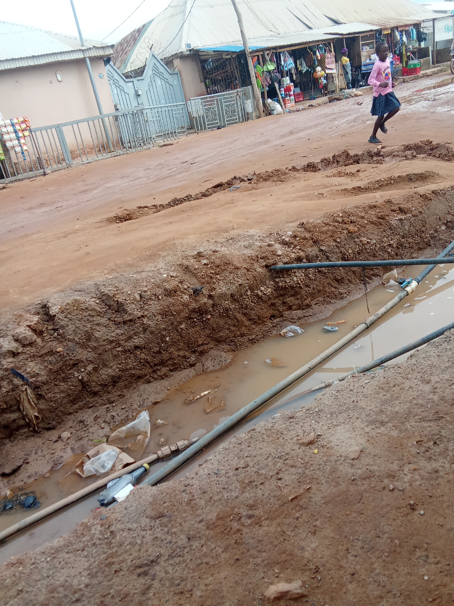 In Kagini, Nigeria, a young girl in a pink long-sleeved shirt and a dark skirt runs along a dirt road. The road is uneven, with a large, muddy trench running alongside it, filled with murky water and debris. Several pipes are submerged in the trench, some appearing to be water pipes. In the background, there are shops with corrugated metal roofs and various goods displayed outside. A metal fence and gate are visible in front of a building to the left of the road.
