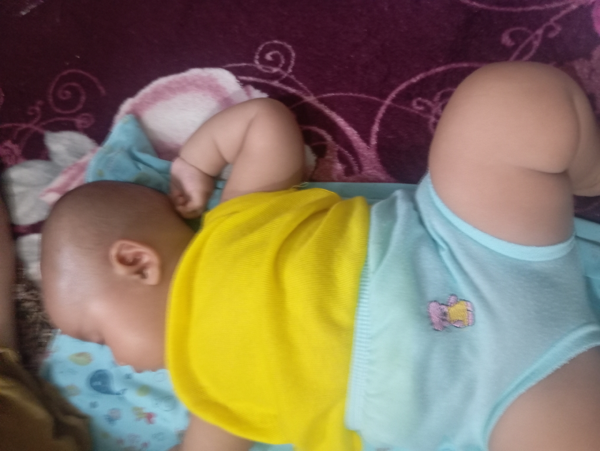 A baby is sleeping on a light blue patterned blanket, which is placed on a dark purple patterned surface. The baby is wearing a bright yellow shirt and light blue shorts with a small embroidered teddy bear. The baby's head is turned to the left, and their right arm is bent, with their hand near their face. The baby's legs are bent, with their feet pointing towards the right. The image is taken from a slightly elevated angle, looking down at the baby. The background shows a blurry depiction of the patterned purple surface, possibly a rug or bedspread. The lighting is soft and diffused, creating a warm atmosphere. The overall scene suggests a peaceful moment of rest for the baby in a domestic setting.