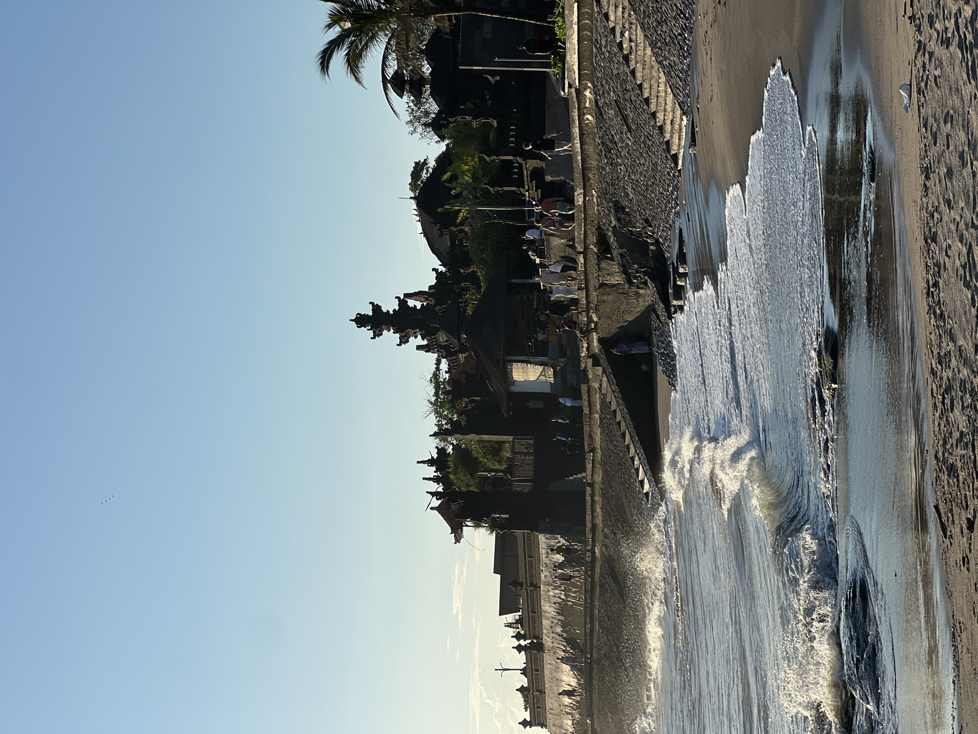 The image depicts the Tanah Lot Temple in North Kuta, Indonesia, situated on a rocky islet off the coast. The temple complex is visible along the coastline, featuring tiered structures and traditional Balinese architecture. People are present on the beach and along the path leading to the temple. Waves are crashing against the shore, indicating a coastal environment. The sky is clear with a light blue hue. The sand is wet and reflects the sunlight.