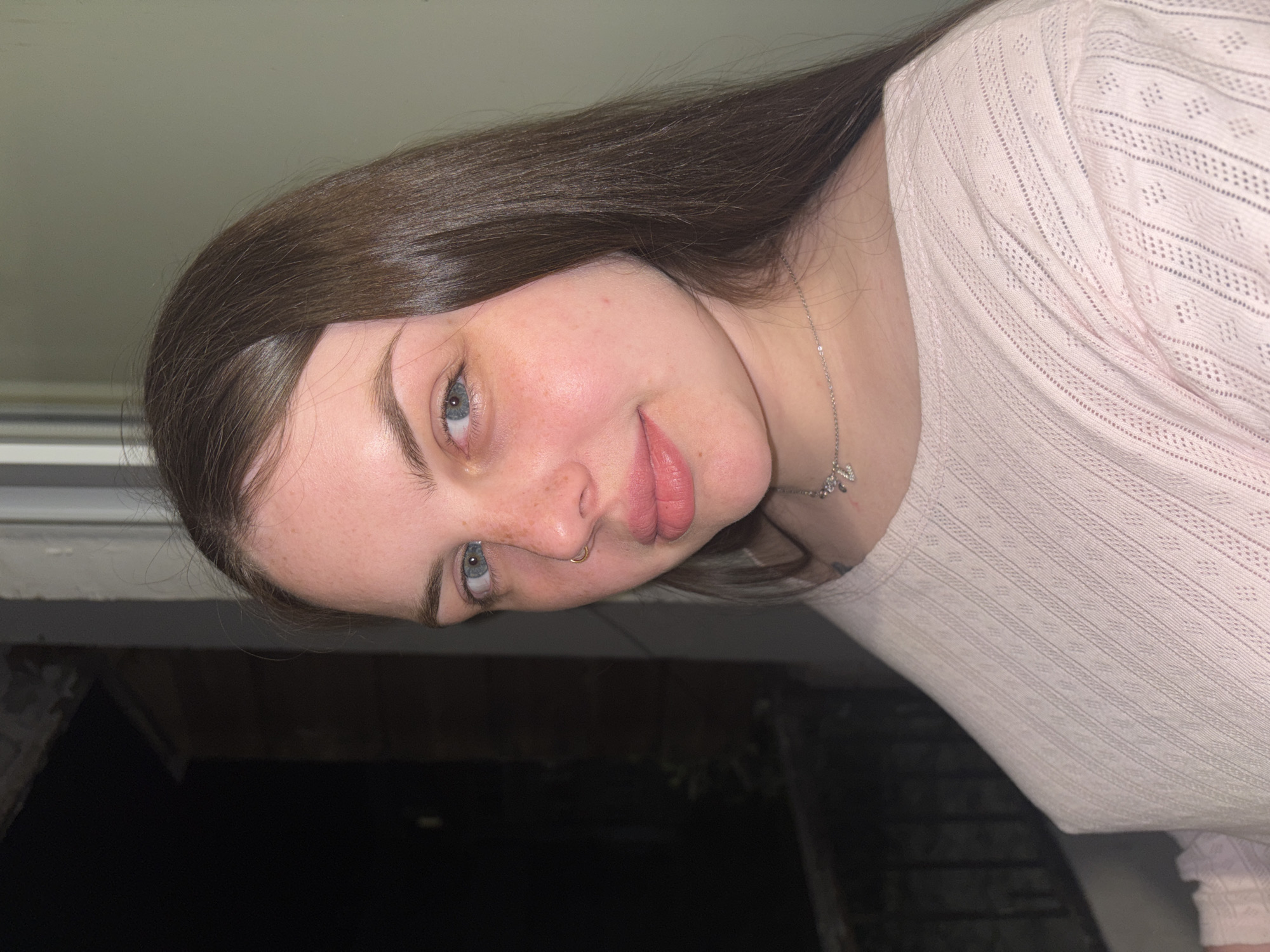 A young woman with long, straight, dark brown hair is captured in a close-up, head-and-shoulders portrait. She has fair skin, light blue-grey eyes, and a scattering of freckles across her nose and cheeks. Her lips are full, and she wears a small gold hoop nose ring in her right nostril. She is looking directly at the camera with a slight, gentle smile or smirk, and her head is subtly tilted to her right.
She is wearing a light pink top with a delicate, textured pattern, possibly knitted or featuring eyelet details. A thin silver necklace with a small, intricate pendant, which appears to be a butterfly or abstract design, is visible around her neck.
The lighting in the image is somewhat dim. The upper left portion of the background is a light, possibly off-white or pale green, wall or ceiling, while the lower right background is very dark, suggesting a low-light indoor setting or nighttime outdoors. There is no visible text in the image. The overall impression is candid and intimate.