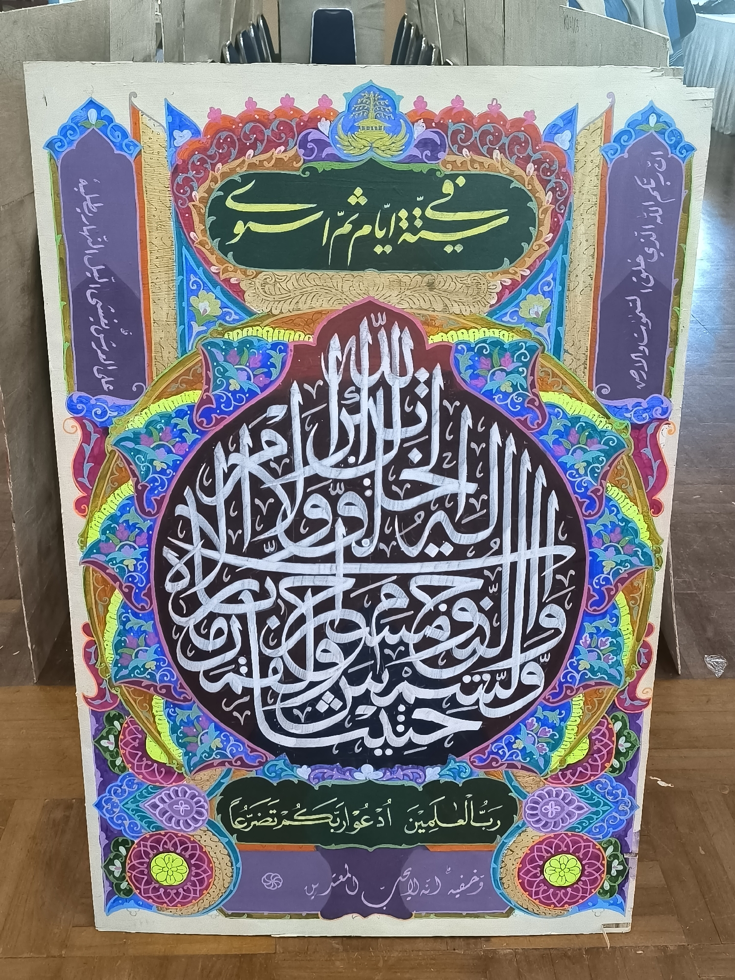 The image shows a colorful Islamic calligraphy artwork displayed on a wooden floor, likely in a public space such as a mosque, community center, or an exhibition hall in the city of Minasa Te'ne, Indonesia. The artwork is painted on a rectangular canvas with intricate designs and Arabic script in white, yellow, and green against a black, green, and dark purple background. The calligraphy appears to be a religious text or prayer. The artwork is framed by a vibrant border with floral and geometric patterns in shades of blue, purple, pink, orange, and yellow. The background of the image reveals the environment where the artwork is displayed. We can see wooden floorboards and the lower parts of what appears to be wooden furniture or partitions, suggesting an indoor setting. There are no people visible in the frame. The lighting appears to be artificial, typical of an indoor event or display.