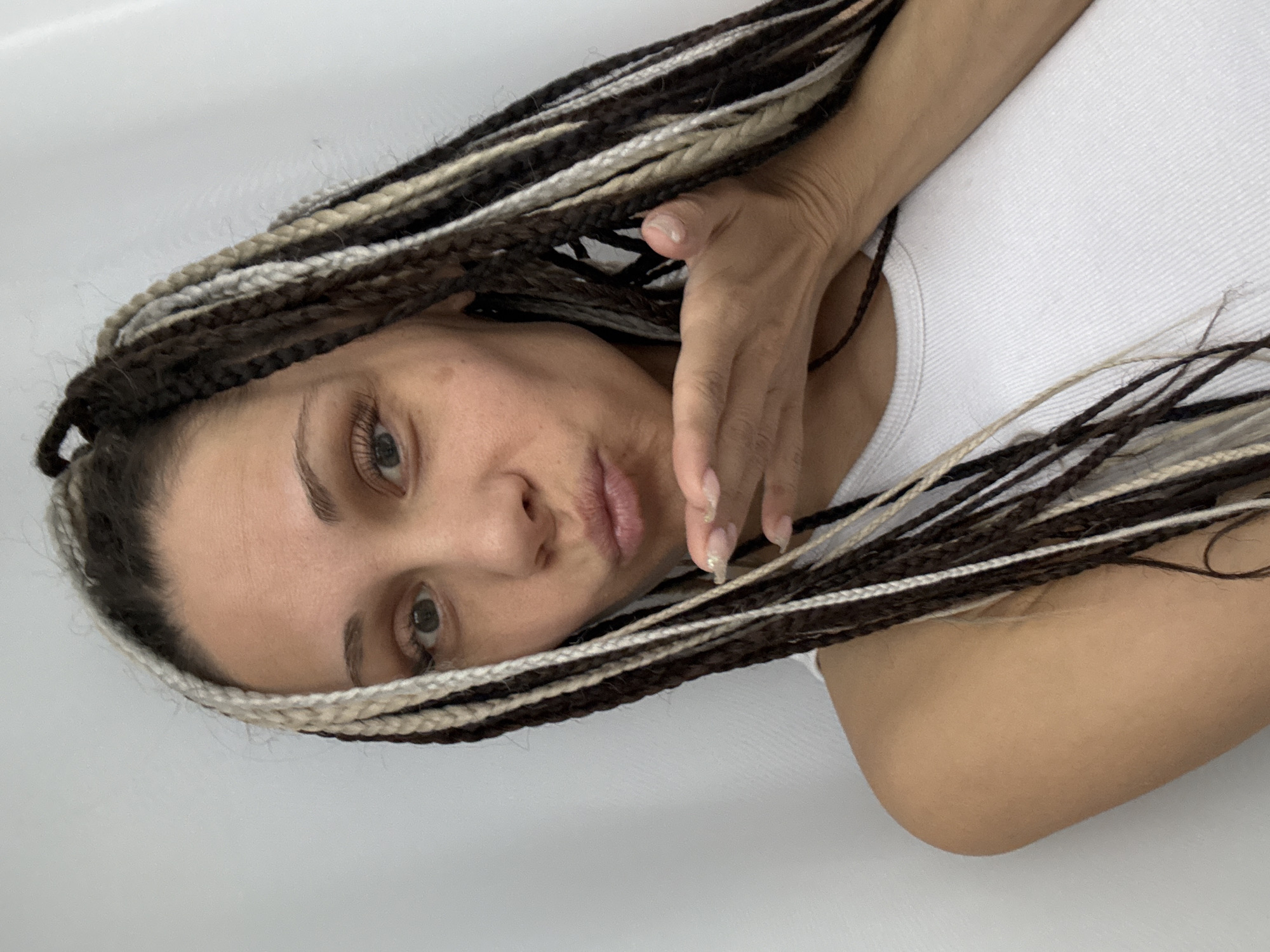 A person with braided hair is lying on their side against a white surface. The braids are a mix of dark brown and blonde colors. The person is wearing a white tank top. Their hand is near their face. The location is not identifiable as Rusanovo, Russia from the image.