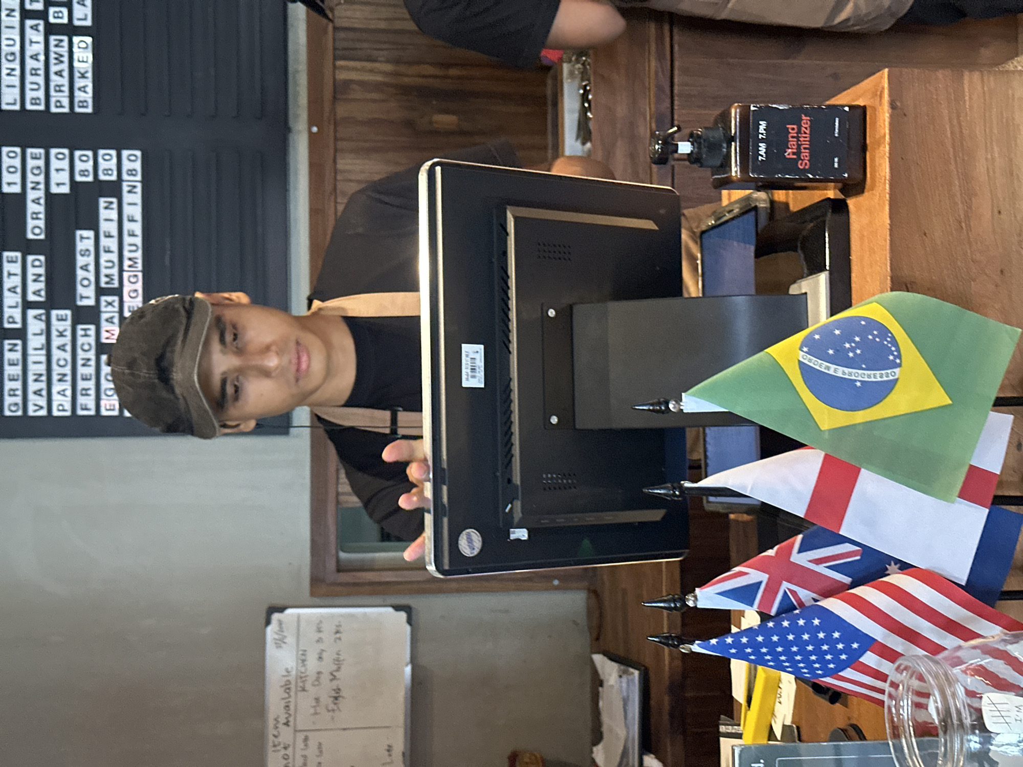 The image shows a young man, likely an employee, standing behind a point-of-sale terminal with his hands on the screen. He is wearing a black t-shirt and a dark cap. In front of the terminal, a display board with menu items and prices is visible. Several small flags, including the flags of the United States, Australia, England, and Brazil, are placed on the counter. To the right of the terminal is a bottle of hand sanitizer. The background features wooden paneling and a gray wall. A whiteboard with handwritten notes is to the left of the employee. The overall scene suggests a cafe or restaurant in North Kuta, Indonesia.