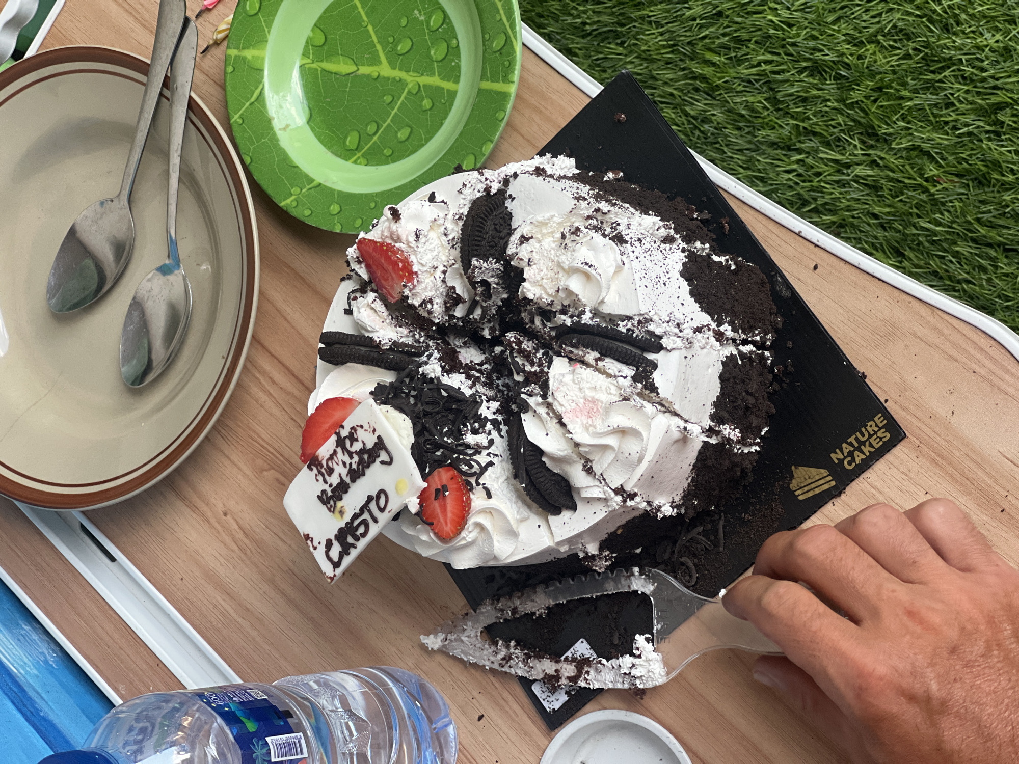 A partially eaten chocolate cake with white frosting and Oreo cookies is on a black cardboard base with "NATURE CAKES" written in gold. The cake has strawberries and a white rectangular topper that reads "Happy Birthday CRISTO". A hand is holding a clear plastic cake server, and a slice of cake has been removed and is resting on the server. The cake is on a light brown wooden table next to a clear plastic water bottle. In the background, there is a beige plate with two spoons and a green decorative plate with leaf imprints and water droplets. The scene is outdoors, with a grassy area visible to the right of the table. The presence of the "NATURE CAKES" branding suggests the cake was purchased from a bakery in Denpasar, Indonesia.