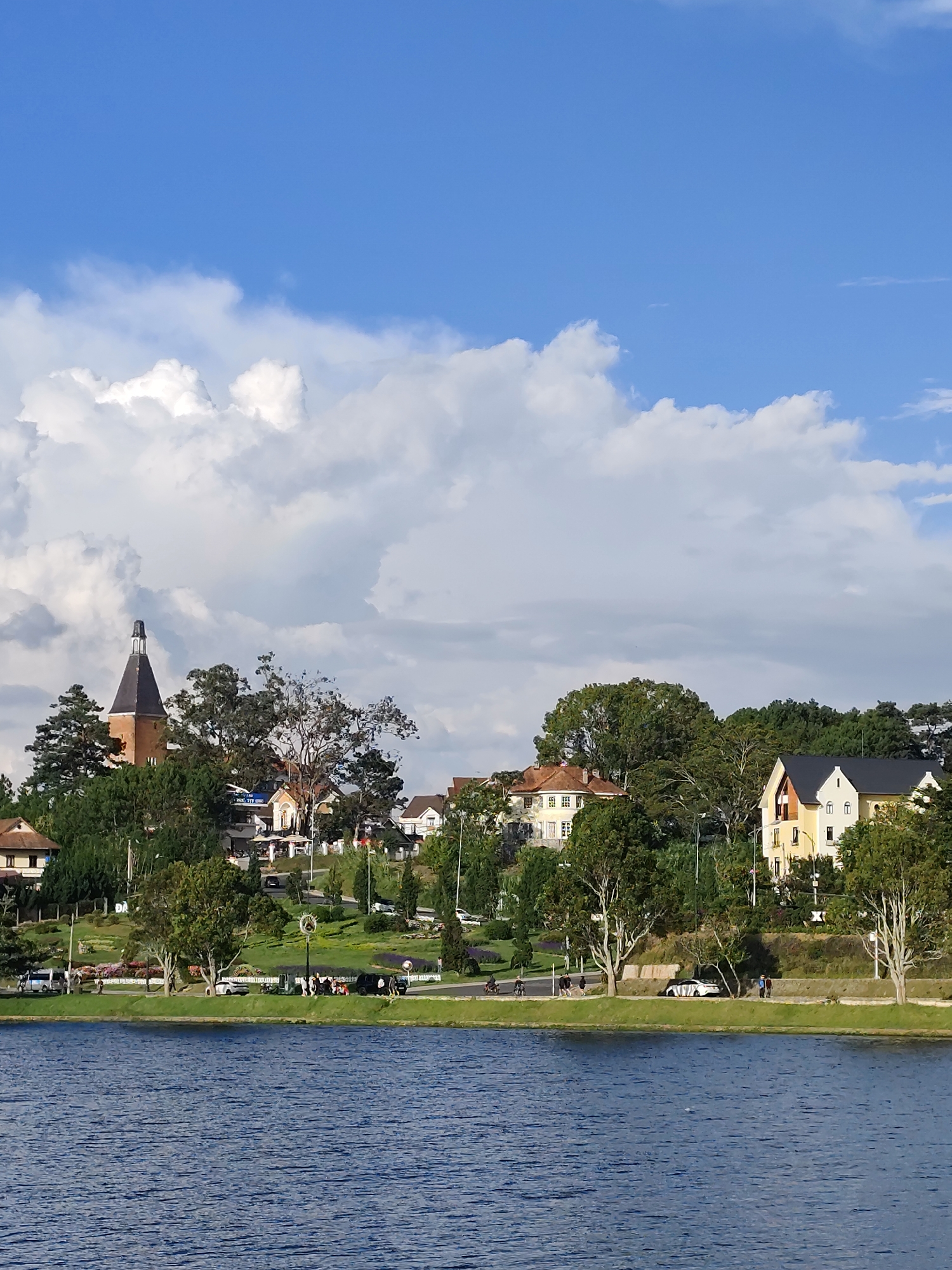 The image depicts Xuan Huong Lake in Da Lat, Vietnam, on a sunny day with scattered white clouds against a clear blue sky. The foreground is dominated by the deep blue water of the lake, with ripples indicating a gentle breeze. Along the far bank, a scenic landscape unfolds. A road runs parallel to the lake, with vehicles, including cars and motorcycles, and several individuals visible. Beyond the road, a gently sloping hill is covered in lush green grass and meticulously maintained flower beds, adding vibrant splashes of color. A row of trees lines the edge of the grassy area, separating it from a cluster of European-style houses and buildings further up the slope. Notably, a prominent building with a distinctive, dark conical steeple stands out on the left side of the frame, suggesting a historical or religious structure. The architecture of the houses varies, with some featuring yellow facades and dark roofs, while others have lighter colors. The overall scene conveys a sense of peaceful coexistence between natural beauty and urban development.