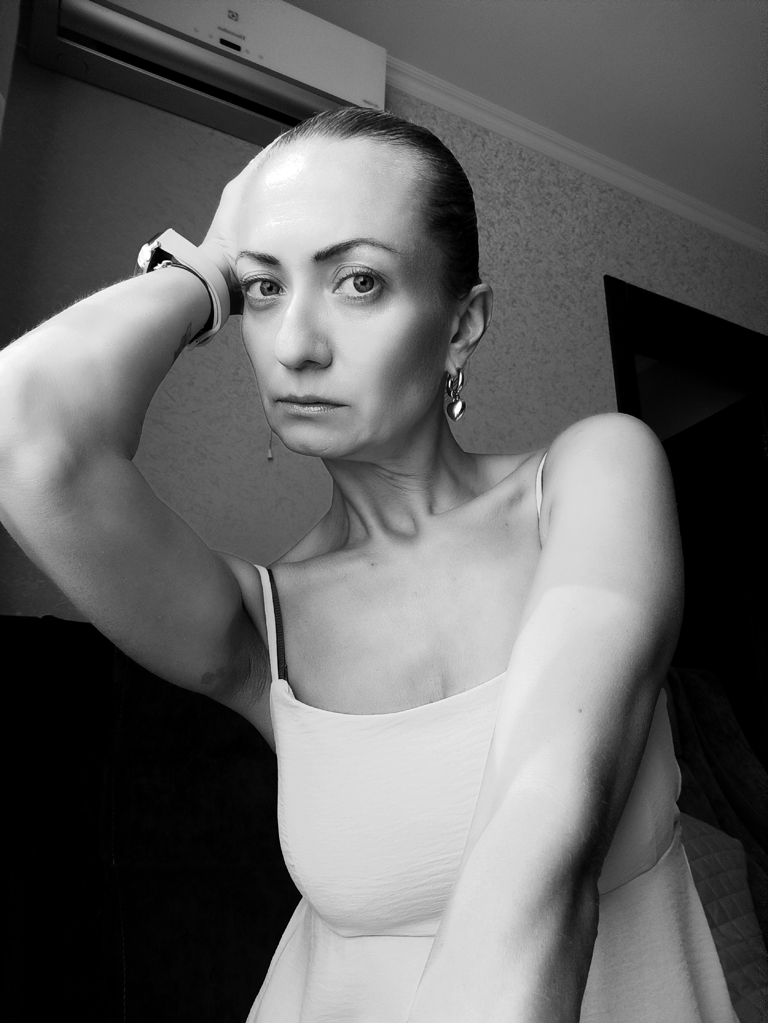 A black and white, medium shot photograph shows a woman's head and upper body. She is looking directly at the camera with a serious expression. Her hair is pulled back from her face. She is wearing a white, sleeveless top and a watch on her left wrist. Her left arm is bent, with her hand resting on her head. There are earrings visible on her ears. The background appears to be an indoor setting with a wall and what looks like an air conditioner unit above her head. The lighting creates strong shadows, particularly on the right side of her face and body. The location is not specified in the image or context provided.