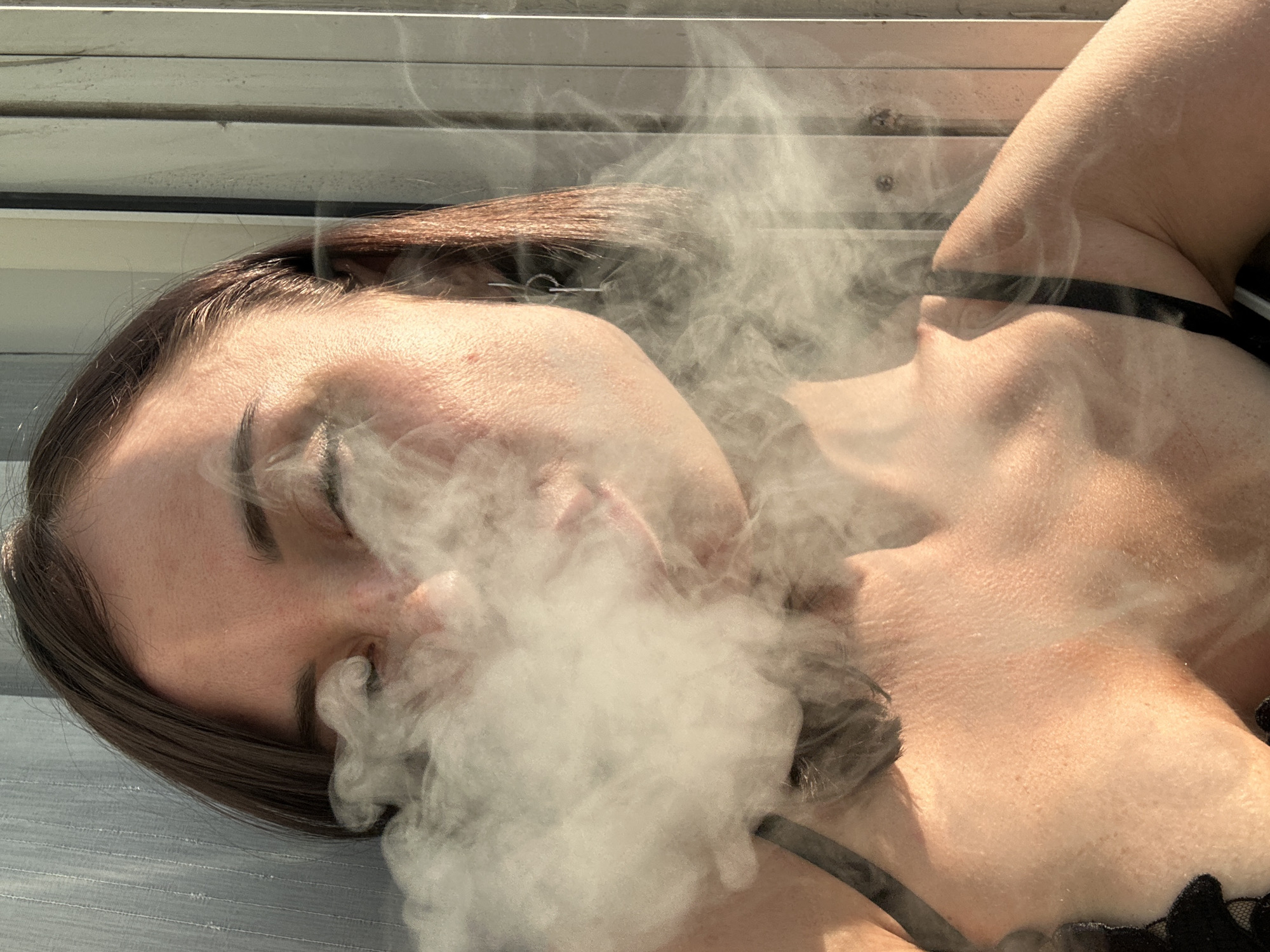 The image shows a close-up, waist-up shot of a young woman with brown hair and closed eyes, exhaling a large cloud of vapor. She is wearing a black bra. The lighting suggests it is daytime, with sunlight casting shadows. The background appears to be an outdoor setting with vertical metallic panels. There are no discernible landmarks that place the image specifically in Nha Trang Ward, Vietnam.