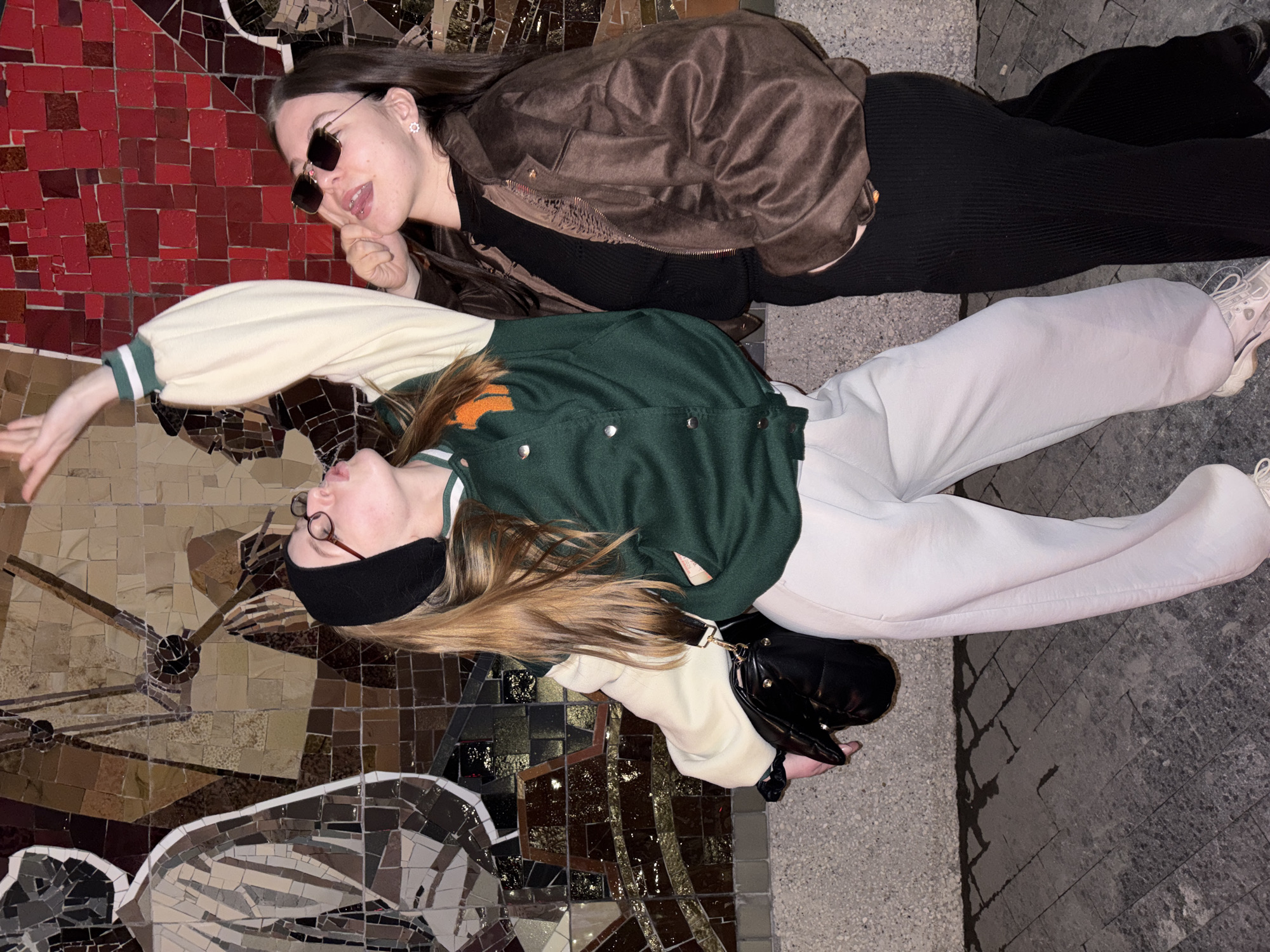 Two women are lying next to each other on a tile mosaic of an image or design. The mosaic appears to be installed on a wall or floor. One woman, wearing sunglasses and a brown jacket, is closer to the top of the image. The second woman is wearing a black eye mask, a green jacket with cream-colored sleeves, and light-colored pants. The women are lying on a stone or concrete surface. The location may be Perm, Russia.