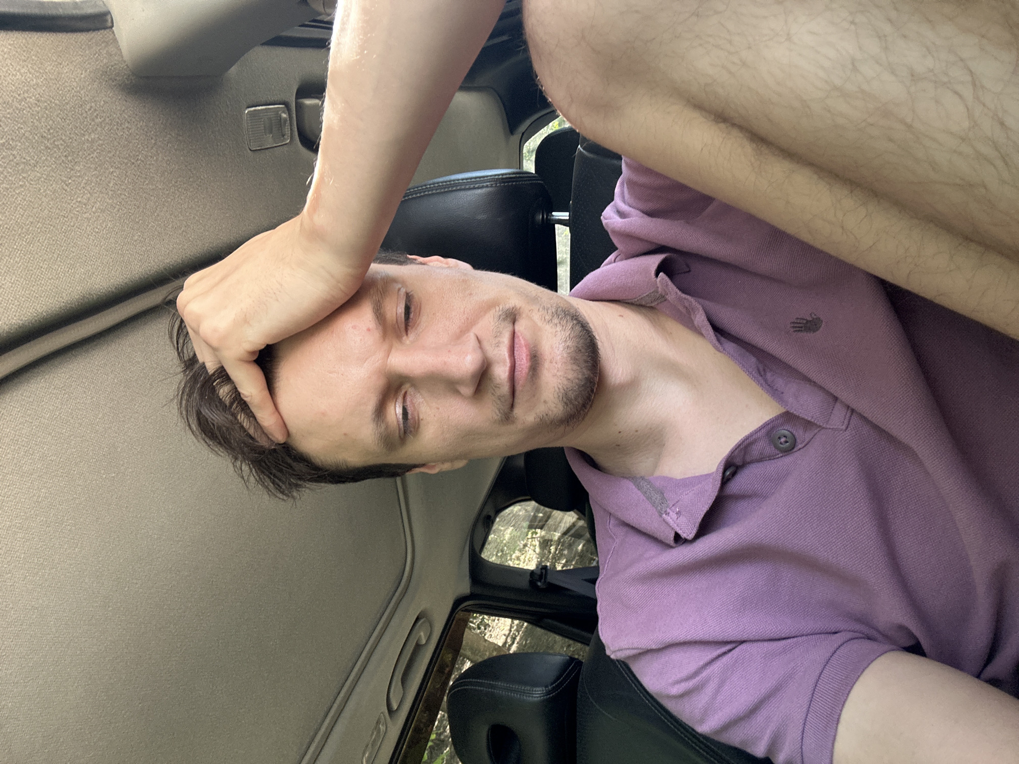 A man is sitting in the passenger seat of a car, holding his hand to his forehead. He is wearing a purple polo shirt. The car appears to be parked on a street in Makiv, Ukraine, as indicated by the surrounding vegetation and the car's interior. The man's expression suggests he may be tired or stressed.