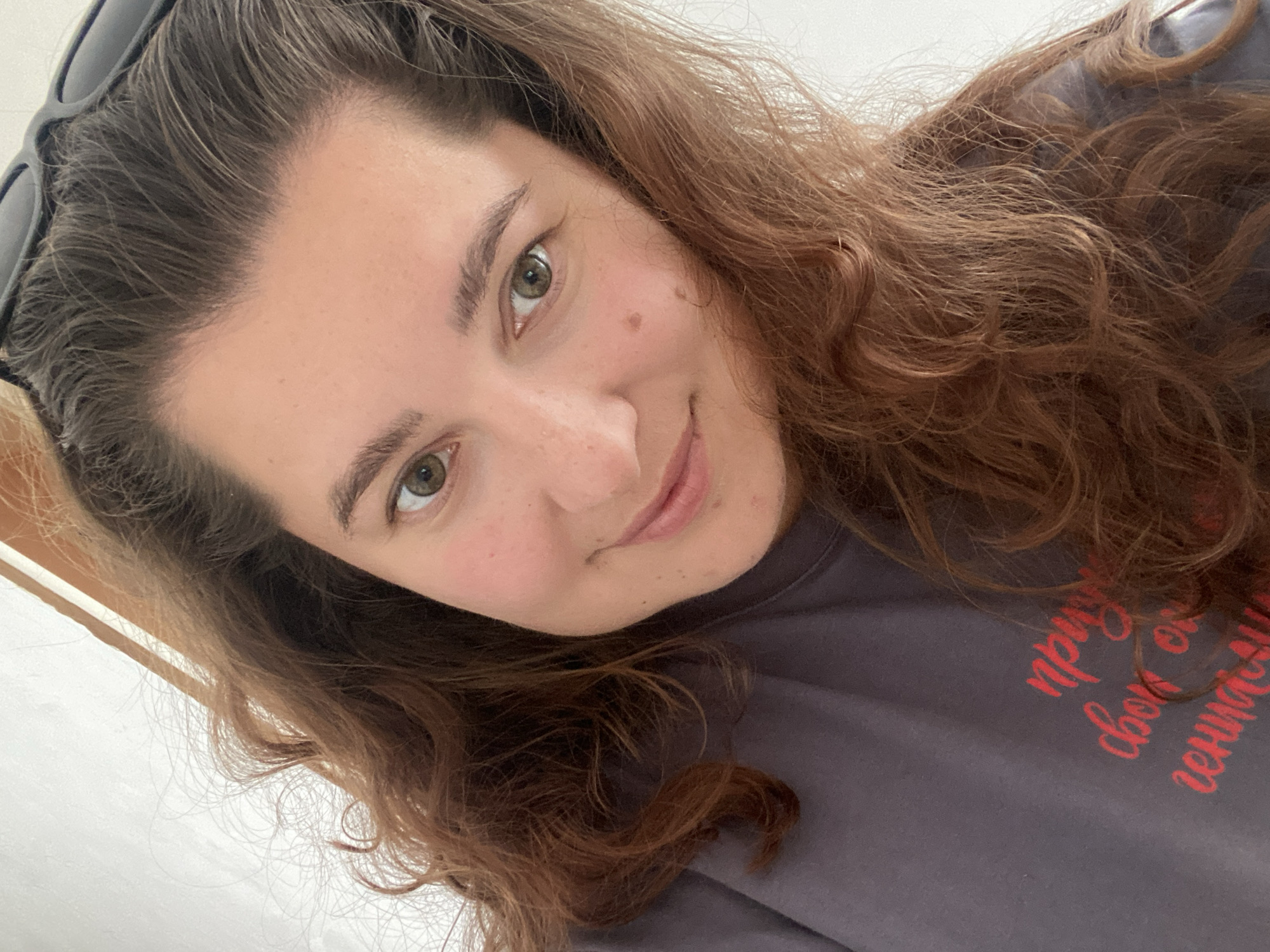 A close-up, selfie-style, slightly angled indoor shot shows a young woman with long, wavy, chestnut-brown hair. She has fair skin with a smattering of freckles, green eyes, and a mole on her right cheek. She is wearing a dark gray t-shirt with red text printed on it, which is partially obscured by her hair. The text appears to be in Cyrillic script. A pair of black sunglasses are perched on top of her head. The background consists of a plain, light-colored wall and a wooden frame, suggesting an indoor setting. There are no discernible landmarks that would indicate the city of Perm, Russia.