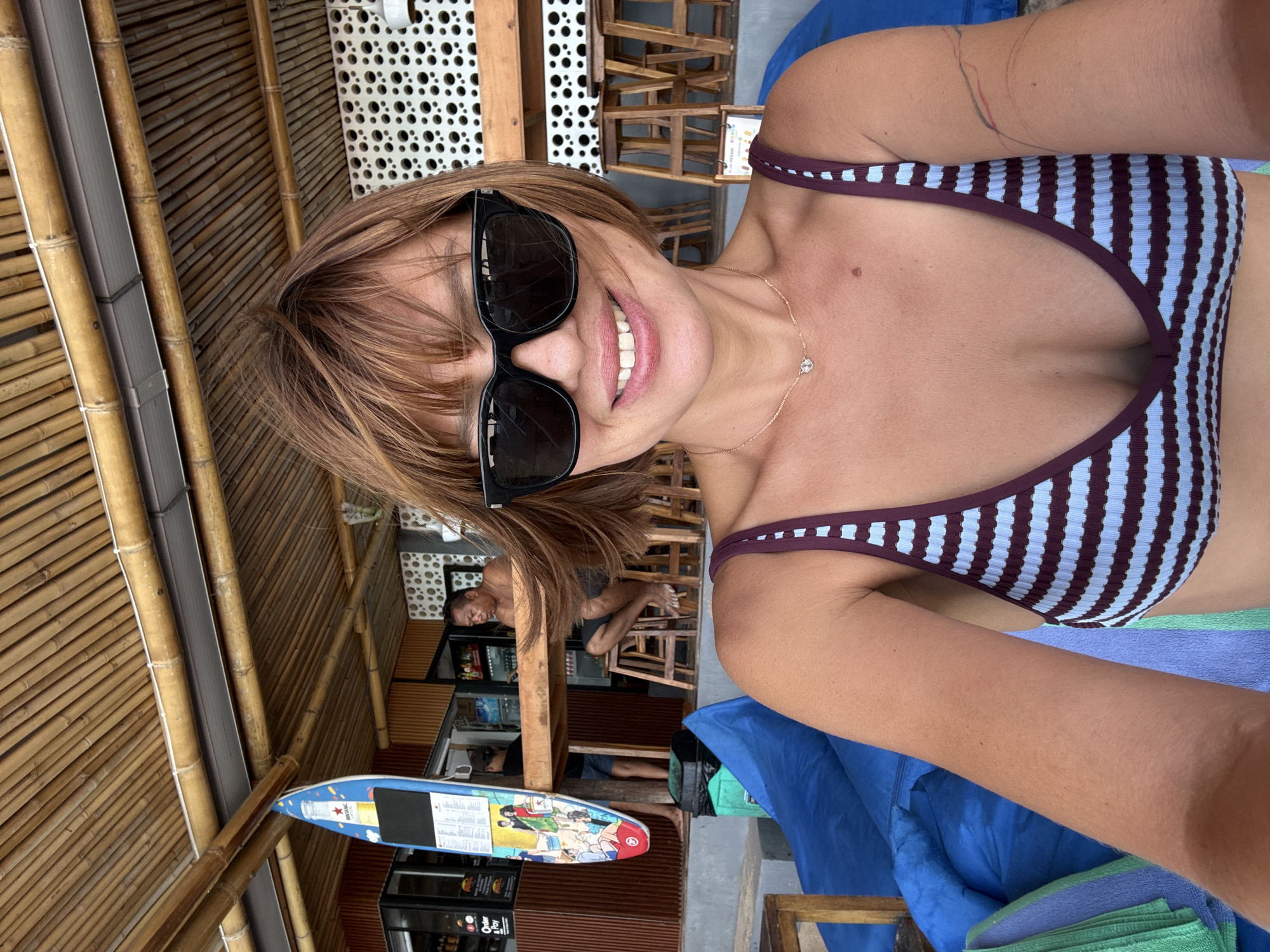 A woman is taking a selfie in a semi-outdoor setting, likely a beach bar or cafe. She is smiling broadly, wearing large dark sunglasses and a striped bikini top in light blue and dark reddish-brown. Her light brown hair with bangs frames her face, and a delicate gold necklace with a small pendant is visible. A faint, colorful linear mark or tattoo is on her upper left arm.
The background reveals a rustic establishment featuring walls and ceiling made of bamboo slats. Several wooden chairs are arranged, suggesting seating for patrons. In the mid-ground, a man with dark hair is seated on a chair, partially obscured by the woman's hair and sunglasses. Another person is partially visible further back near what appears to be a refrigerator or cooler unit. A distinctive blue surfboard-shaped menu board stands upright, and a white perforated panel is visible overhead. The woman is seated, with blue and green towels or fabric beneath her.
The activity is a casual selfie, capturing a moment of apparent relaxation and happiness. The bright lighting indicates it is daytime. The bamboo architecture and the presence of a menu board suggest a tropical or beachside location, consistent with Kuta Selatan, Indonesia.
Visible text includes the "Heineken" logo on the blue surfboard menu board, along with "Order & Pay" at the bottom. Other smaller text on the board is illegible.