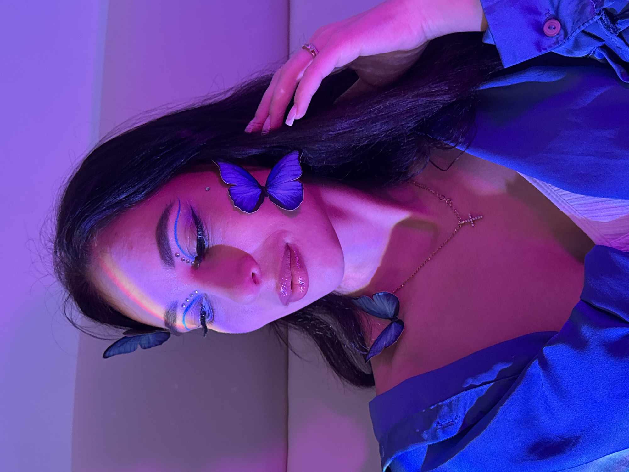 A portrait of a woman with dark hair is illuminated by pink and blue lighting. She is lying down, with her head tilted to the left. Three blue butterfly decorations are present: one on her forehead near her right eye, one near her left ear, and one on her collarbone. She has blue eyeliner and small rhinestones decorating her eyes. She wears a necklace with a cross pendant and a blue top. A ring is visible on her left hand. The background appears to be a plain wall. The location cannot be determined from the image.