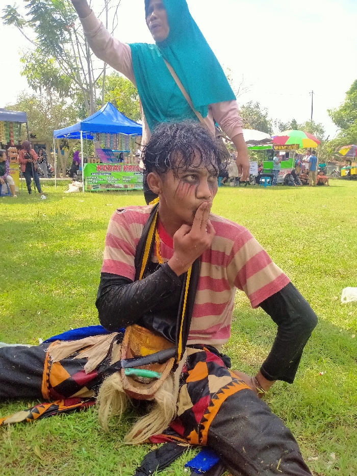 A person in costume sits on green grass. Dark makeup streaks down their face, and they wear a pink and white striped shirt with black sleeves. A decorative piece with a wooden mask rests on their lap. Behind the person, an adult in a turquoise hijab raises their arm. In the background, tents and people suggest a festival or outdoor event. The overall scene is captured in a casual, natural setting.