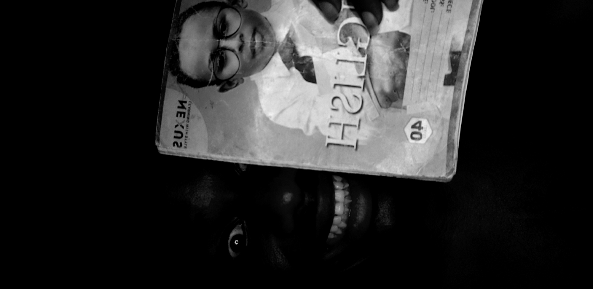 A black and white photograph captures a dark-skinned person partially obscured by a magazine cover, set against a completely dark background in Narayi, Nigeria. The magazine cover is tilted, revealing only one wide-open eye and a broad, toothy smile of the person holding it in the lower part of the frame.
The magazine cover itself features a close-up image of another person wearing round glasses, looking directly at the viewer with a subtle smile. Several pieces of text are visible on the cover: "NEXUS" is printed in a horizontally mirrored fashion on the left side, with "LEARNING WITH SLATE" appearing underneath it, also mirrored. The word "HIGH" is prominently displayed and rotated, with a stylized or partially mirrored appearance for some letters. A hexagonal or octagonal emblem containing the number "40" is visible on the right side of the cover. The overall mood conveyed by the visible smile is cheerful.