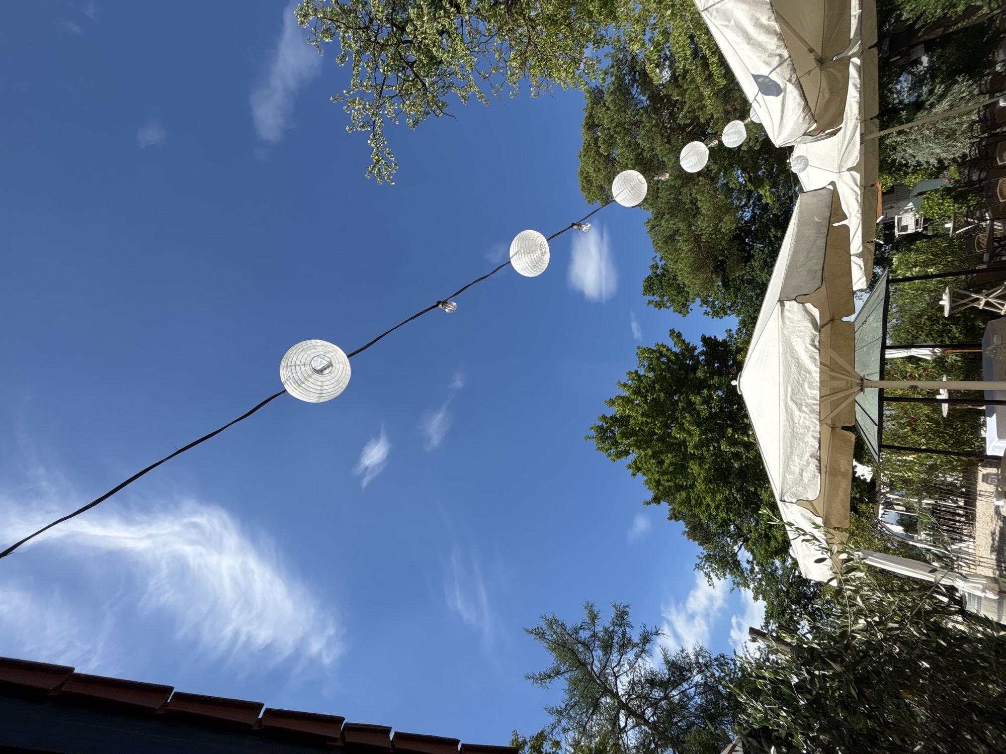 The image captures a bright, sunny day with a clear blue sky dotted with wispy white clouds. A string of white paper lanterns, illuminated from within, hangs diagonally across the frame, strung on a black wire. These lanterns are interspersed with small, clear light bulbs.
The scene appears to be outdoors, possibly in a garden or patio area. Lush green trees with dense foliage frame the upper parts of the image. In the foreground, the edge of a terracotta tiled roof is visible. Part of a white umbrella or canopy is also present in the lower right section of the image, suggesting an outdoor seating area, perhaps for a cafe or restaurant. The overall impression is one of a relaxed and festive outdoor atmosphere, suitable for an event or a pleasant day out. The sunlight suggests it is daytime. No people are visible in the image. There is no visible text.