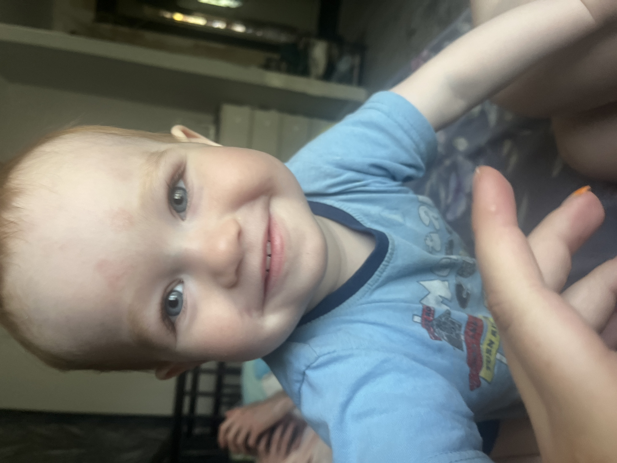 A close-up shot depicts a young child with fair skin and reddish-blond hair. The child is smiling and wearing a blue t-shirt with a graphic print. The child's eyes are blue. A hand with painted nails (color unclear) is visible in the foreground, slightly obscuring the view. The background is blurry, revealing furniture and possible window/window frame. The location cannot be determined based on the image, nor can the city of Chaikovsky, Russia.