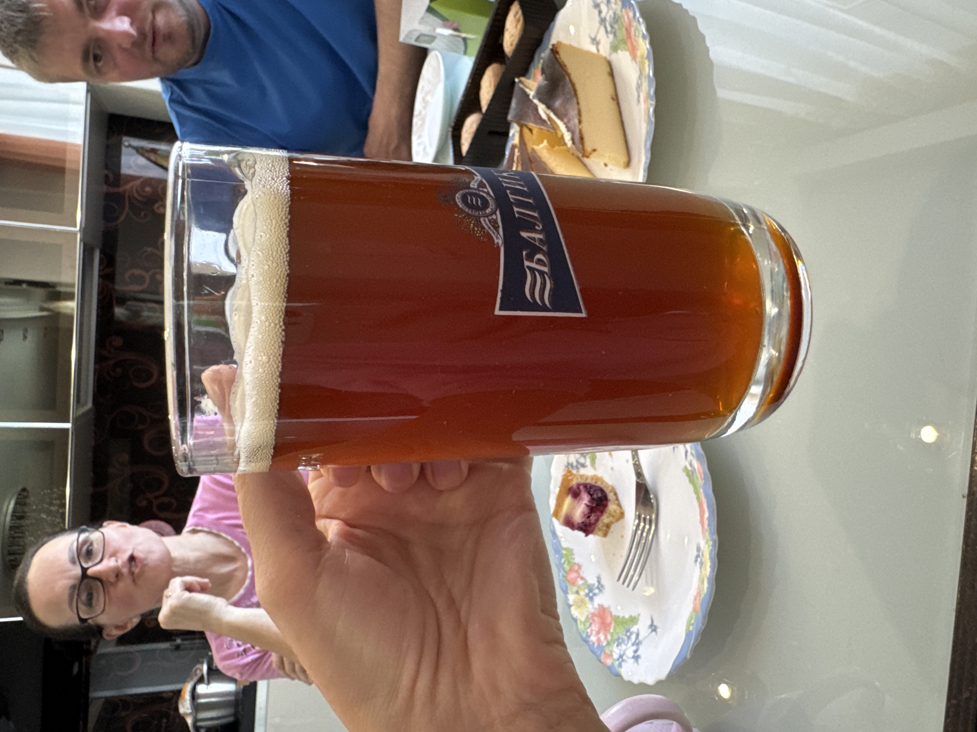 The image shows a partially consumed glass of Baltika beer held in a hand indoors. The beer has a dark amber color and a foamy head. The label on the glass identifies the beer as Baltika, a Russian brand.
In the background, one person is wearing a blue shirt and another person is wearing glasses and a pink shirt. Plates of cake and pastries are on the table near the glass of beer. The setting appears to be a dining area in a home, and there are kitchen cabinets visible in the background.
While it is impossible to definitively determine the exact location is in Saint Petersburg, Russia, based solely on this image, the presence of Baltika beer, a brand originating in that city, strongly suggests a Russian setting.