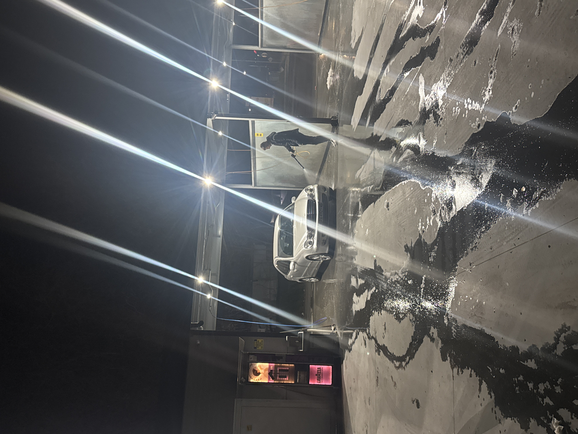 A white car is being power washed at a self-service car wash station in Plovdiv, Bulgaria. A person in dark clothing holds the power washing wand and directs the spray toward the car. The car wash station has multiple bays with overhead lights that cast bright streaks across the image. The ground is wet and streaked with dirt and water. A Lavazza coffee vending machine is visible near the car wash controls.