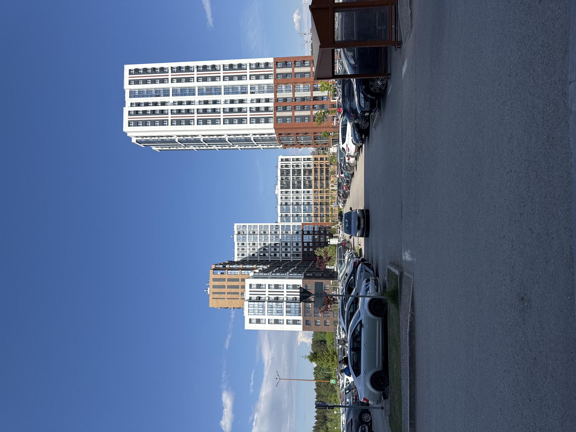 The image shows a view of several modern apartment buildings against a blue sky with some clouds, likely in Perm, Russia. A row of parked cars lines the street. A bus shelter is visible on the right. The photo is rotated 90 degrees counterclockwise.