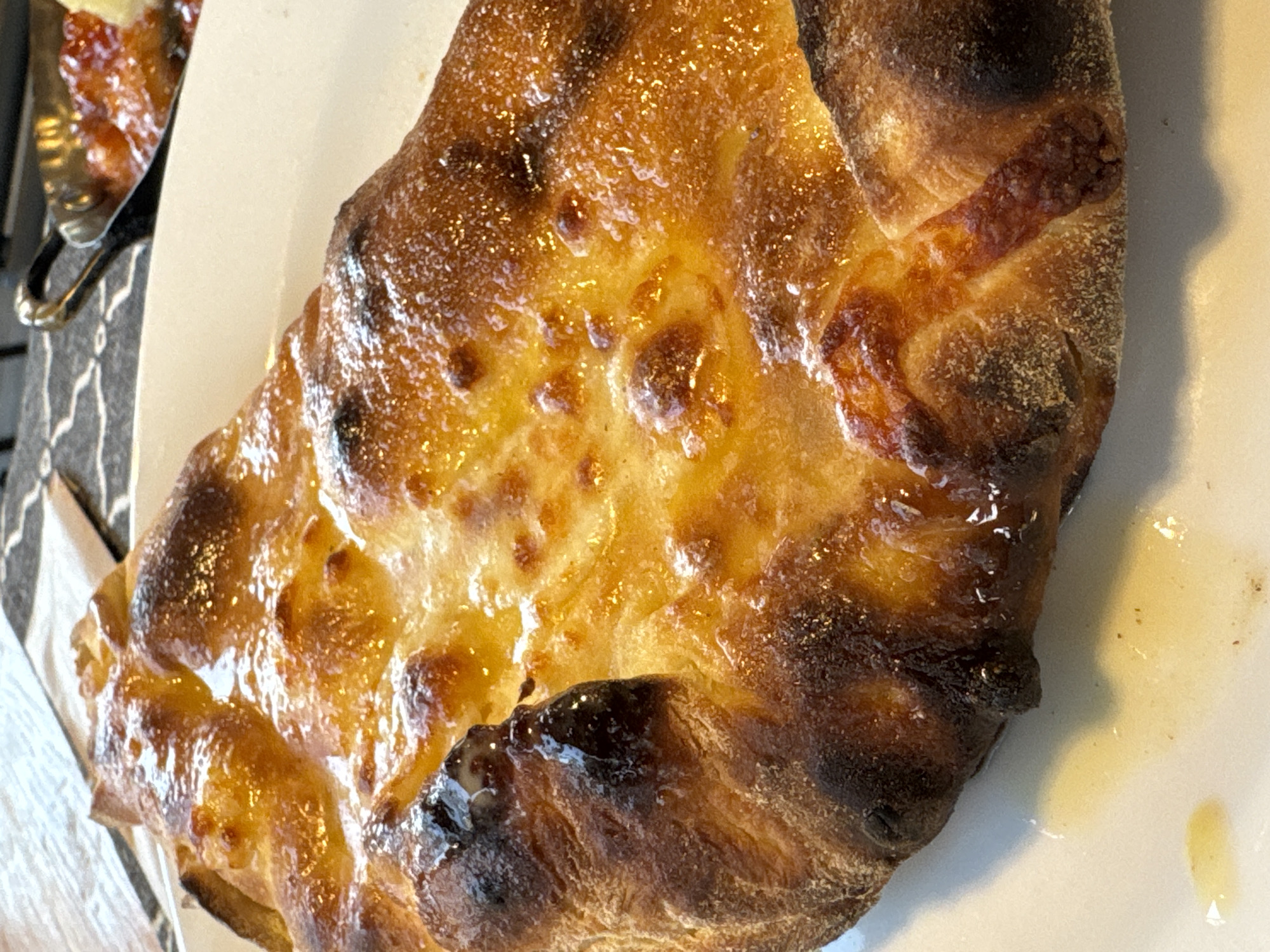 The image shows a close-up of a freshly baked calzone served on a white plate. The calzone has a golden-brown, slightly charred crust with melted cheese oozing from its center. A small dish containing a red sauce and what appears to be a piece of cheese is visible in the background, along with a patterned placemat and a black metal utensil. The scene is likely set in a restaurant in Primorsko, Bulgaria.