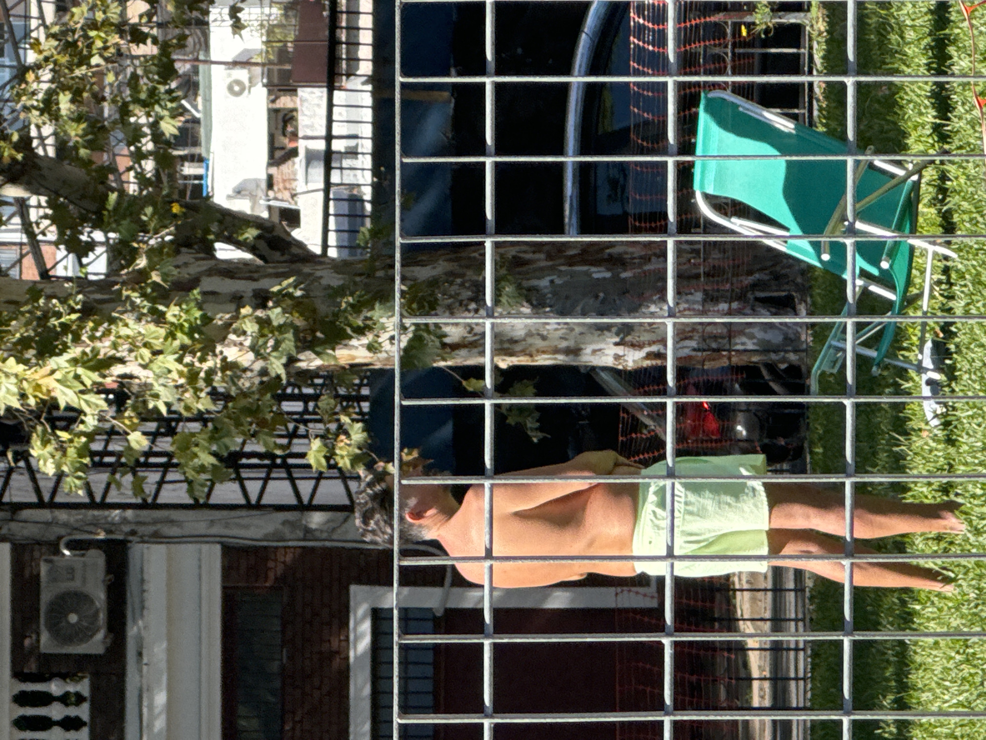 The image presents an outdoor urban scene viewed through a prominent metal grate that spans the entire foreground, creating a grid pattern over the landscape. The photograph appears to be taken from an elevated perspective, looking down.
The central subject is a man lying on his back behind the metal grate, seemingly relaxing or sunbathing. He is shirtless and wears light-colored shorts. His arms are bent, and his legs are extended. To his right, a bright green folding chair is partially visible, positioned in an area with green grass.
In the background, on the left side, are several multi-story buildings with brick and lighter-toned facades. A white air conditioning unit is mounted on one of the building walls. A large tree with abundant green foliage partially obscures these buildings. On the right side, behind the grate, fragments of what appears to be a dark vehicle or structure are visible, along with some orange construction netting.
The scene is brightly illuminated, suggesting clear, sunny daytime conditions. The overall atmosphere appears calm and leisurely. There is no visible text in the image. While the provided context indicates the location is Buenos Aires, Argentina, there are no specific visual cues within the frame to confirm this.