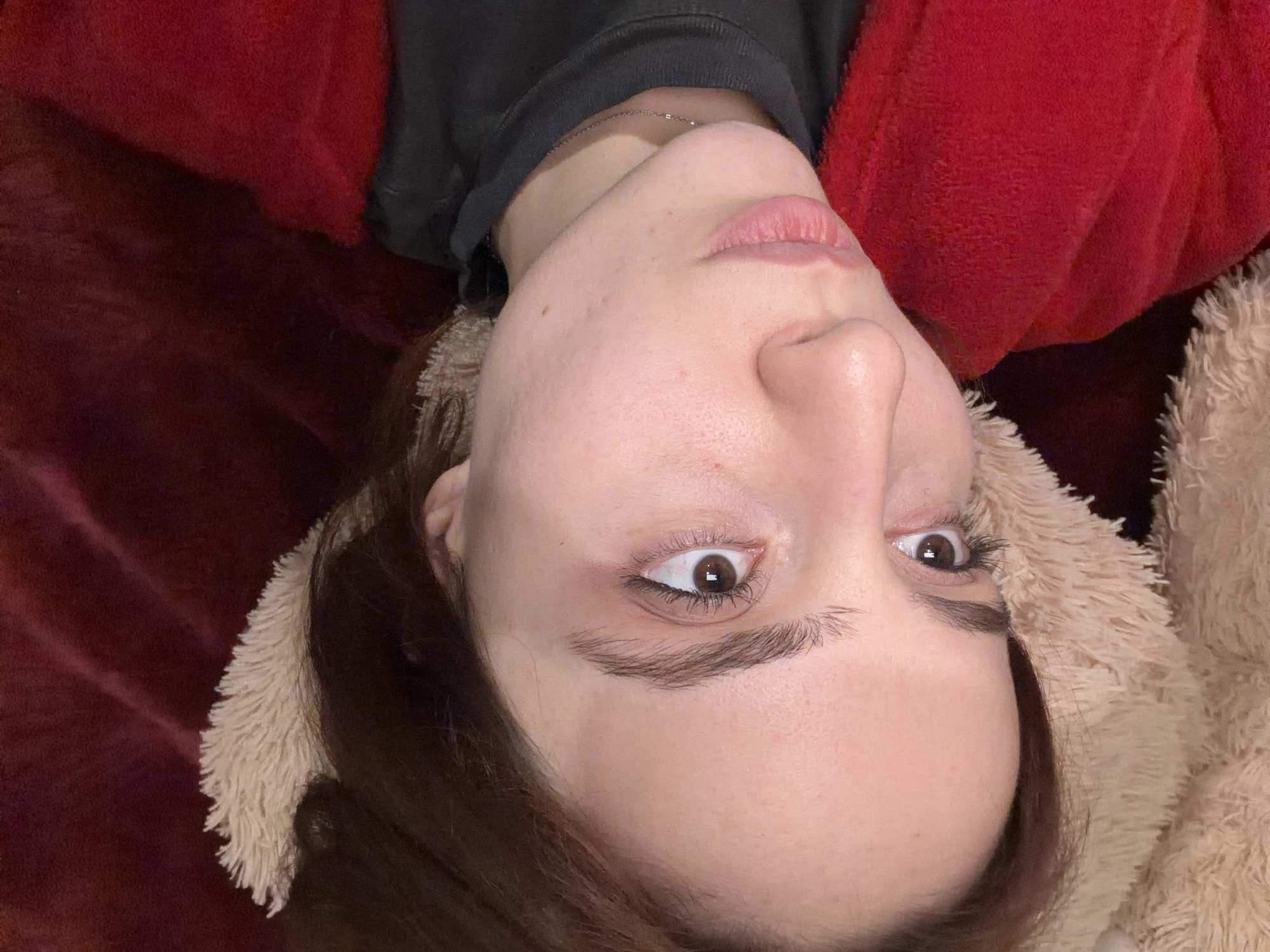 A close-up image depicts a female individual in a reclining position within an indoor, private setting. Her head is slightly tilted, with dark brown hair visible. She has fair skin, prominent brown eyes gazing towards the upper left, defined eyebrows, a slightly upturned nose, and full, naturally pink lips that are slightly parted. A thin silver-toned necklace chain is visible around her neck. She is wearing a dark-colored, high-collared garment underneath a vibrant red, textured, soft outer layer, resembling a robe or blanket. Her head rests against a light beige, fluffy material, while the background consists of a blurred, dark red/burgundy soft fabric. The specific location within the city of Strasbourg, France, cannot be determined from the visual evidence presented in the image.