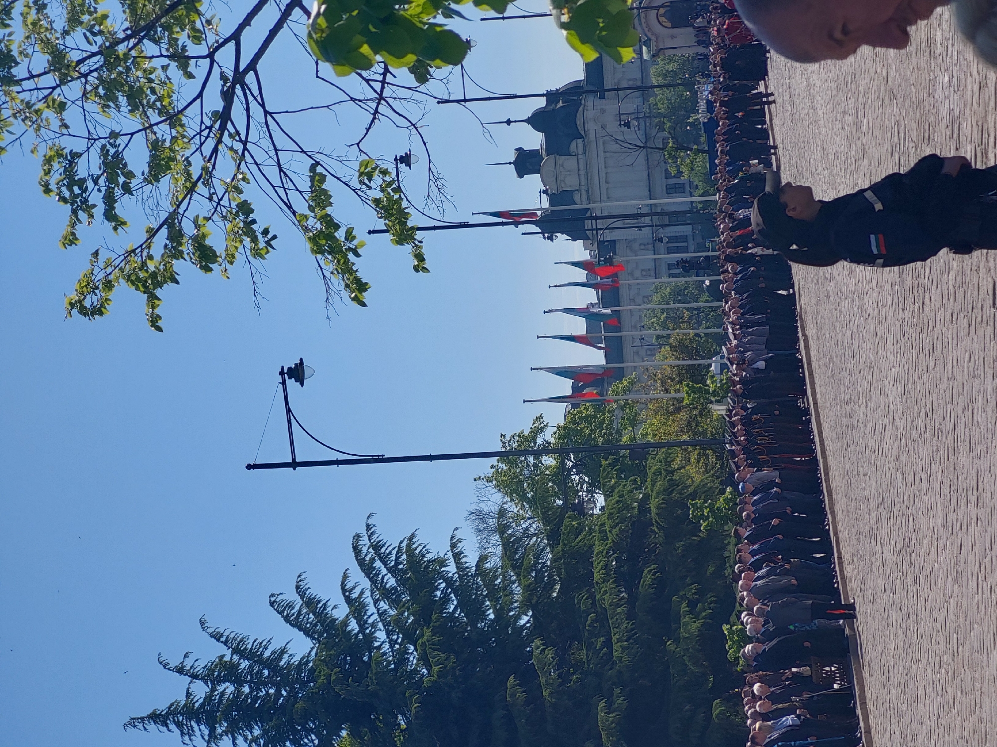 The image shows a row of people standing on a brick walkway in an urban setting. The people appear to be uniformed personnel, possibly military or law enforcement, standing at attention. Behind them are flag poles with flags of red, white, and green stripes. In the background is a large, light-colored building with multiple spires and domes. A person in a dark uniform with red and white stripes on the sleeve is standing on the walkway, looking towards the group. The sky is blue and there are trees in the background. The location appears to be in the city of Unknown, Unknown.