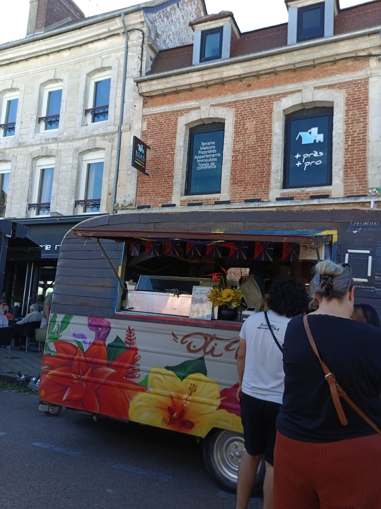 The image depicts a food truck adorned with floral artwork, parked on a street in Le Neubourg, France. In the background, two buildings are visible: a light-colored stone building with multiple windows and a red brick building with framed windows. A sign for "BIAS IMMOBILIER" hangs on the brick building. The food truck has its service window open, with a display of items inside. Several people are standing near the food truck, some appearing to be customers. The street in the foreground is paved and has painted lines. The overall scene suggests a lively street setting, possibly during a market or event.