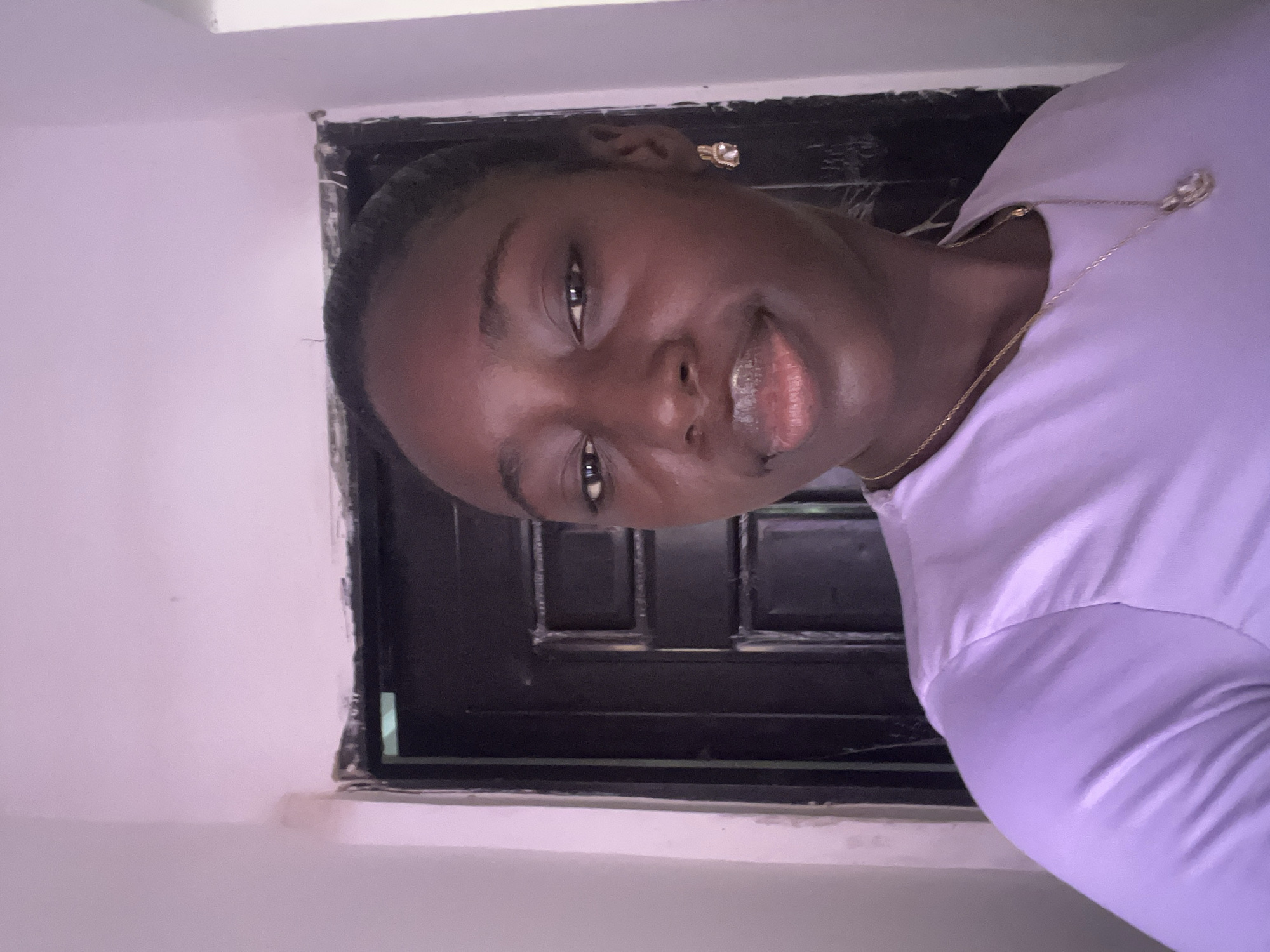 A medium shot presents a dark-skinned woman with short, black hair pulled back from her face. She is wearing a light purple, long-sleeved shirt and a gold necklace and earrings. She is smiling slightly, with a glimpse of her upper teeth visible. Behind her is a black door with panels. The woman and door are set against a light purple or white wall. There is no readily available information to confirm that the image was taken in Abuja, Nigeria, based solely on the visual elements provided. Therefore, I cannot confirm the location.