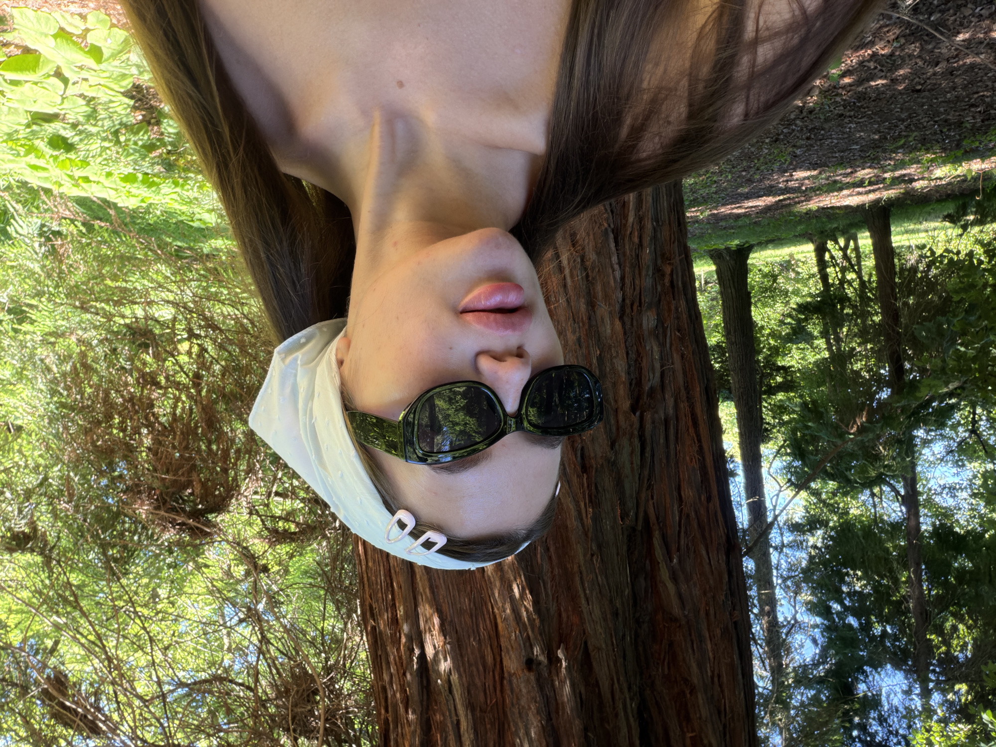 A young woman with long, brown hair is pictured in a wooded area. She is wearing a white headscarf and dark sunglasses. The woman is positioned in the foreground, with her body angled to the left. Behind her, a large tree trunk is visible, and beyond that, a sun-dappled forest floor with green foliage. The location is identified as Sirius, Russia.