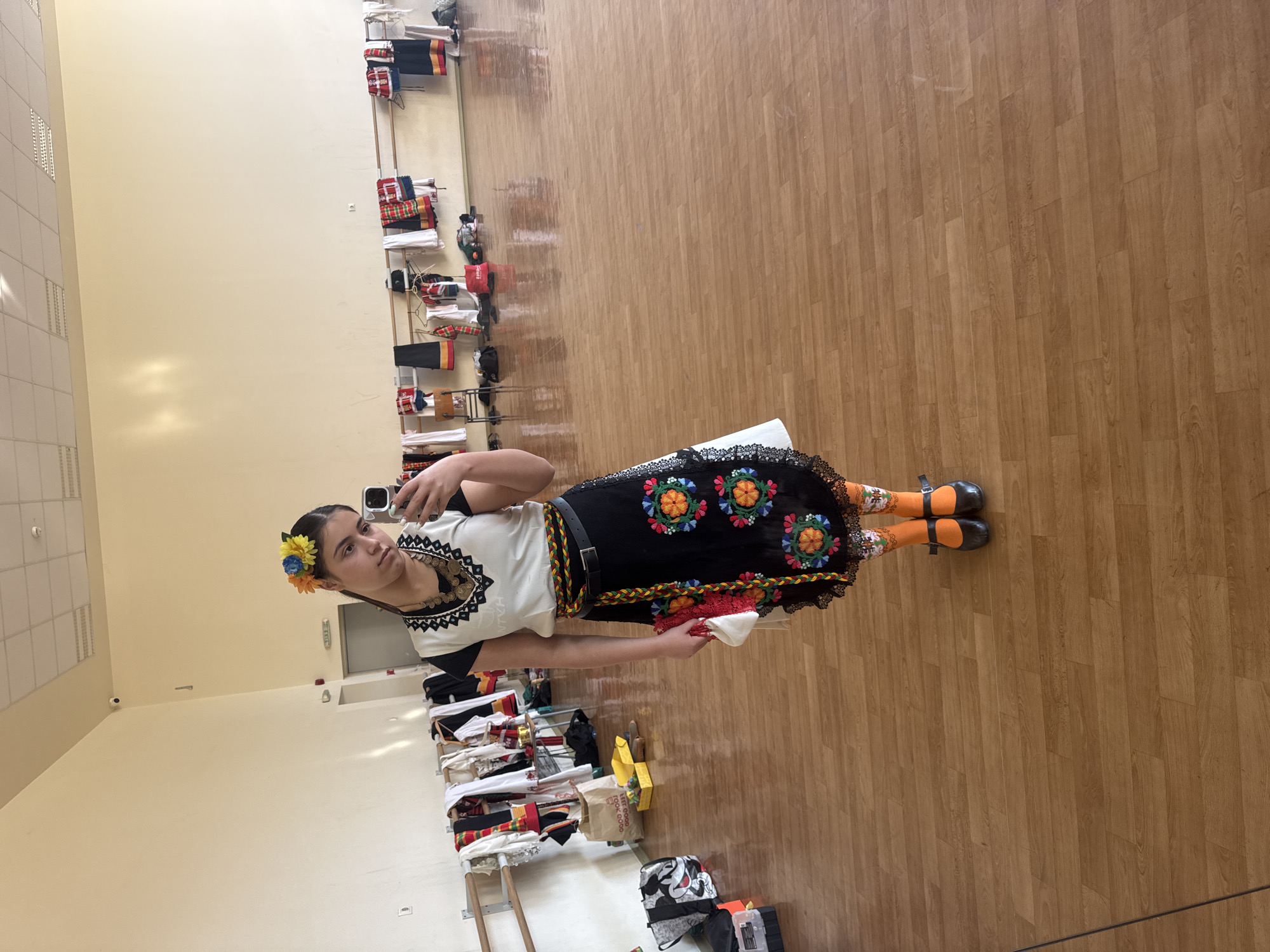 A young woman is standing in a room with light wooden flooring and light yellow walls. She is wearing a traditional folk costume, consisting of a white blouse with black lace trim, a black skirt with colorful embroidered flower designs and a black belt, orange patterned socks, and black shoes with a strap. A yellow and blue flower is clipped into her hair. She is holding a mobile phone in her right hand, presumably taking a selfie. In her left hand, she holds a red and white object. Behind her, along the wall, are several metal racks holding various items of clothing, suggesting a backstage or storage area. The location appears to be indoors, possibly in a cultural center or dance studio.