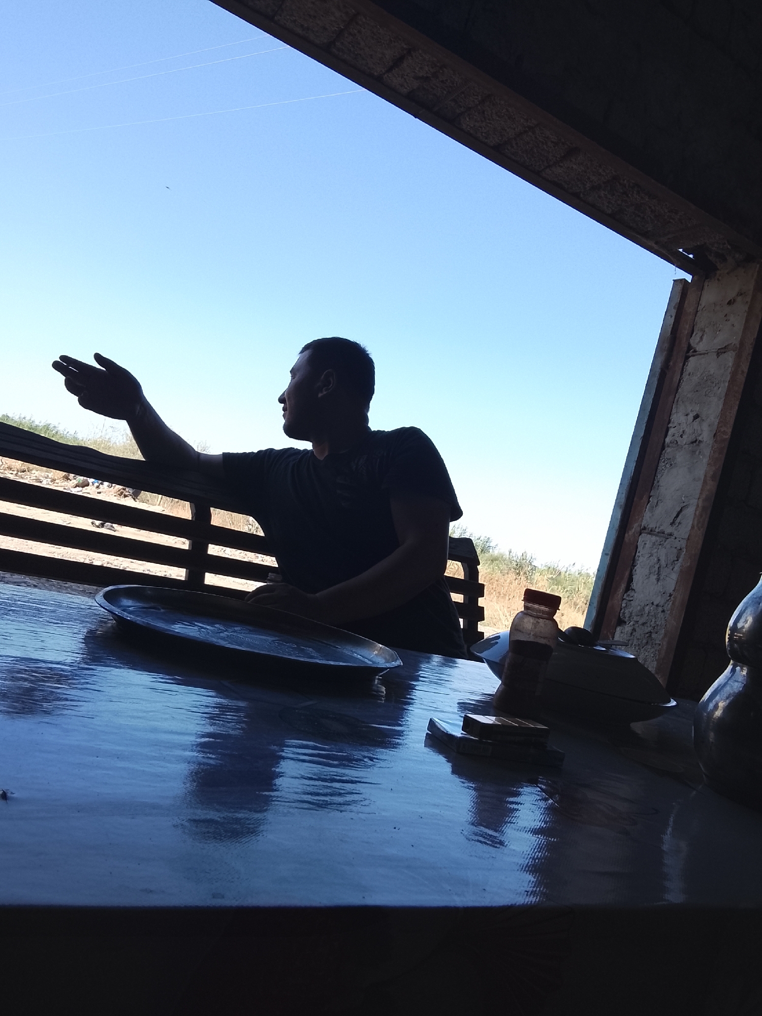 A silhouette of a man sits at a table under a simple structure. He is looking to his left, with his right arm extended and hand open as if gesturing. The table is dark and reflective, with a metal plate, a bottle, and a pack of cigarettes on it. The background shows a clear blue sky and a wooden fence. The setting appears to be outdoors, possibly in a rural or semi-rural area. The image is taken from a low angle, looking upwards towards the sky.