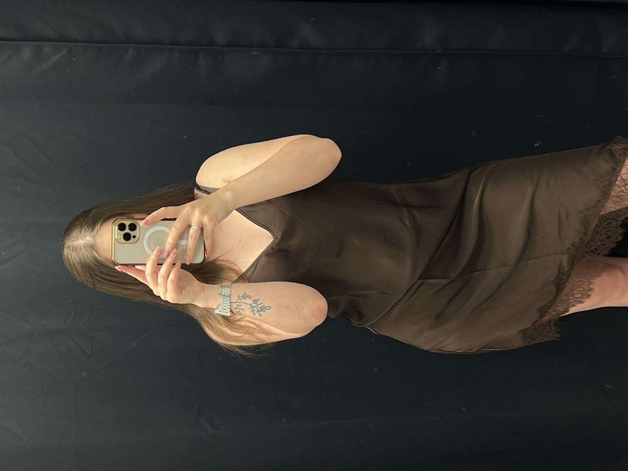 A person with long brown hair is taking a selfie in a mirror. The person is holding a gold-colored iPhone in front of their face and wearing a sleeveless, brown slip dress. The person's left arm is visible, showing a tattoo of a leafy branch and a silver watch. The background appears to be a black wall. There is no visible evidence that suggests the location of Ternopil, Ukraine.