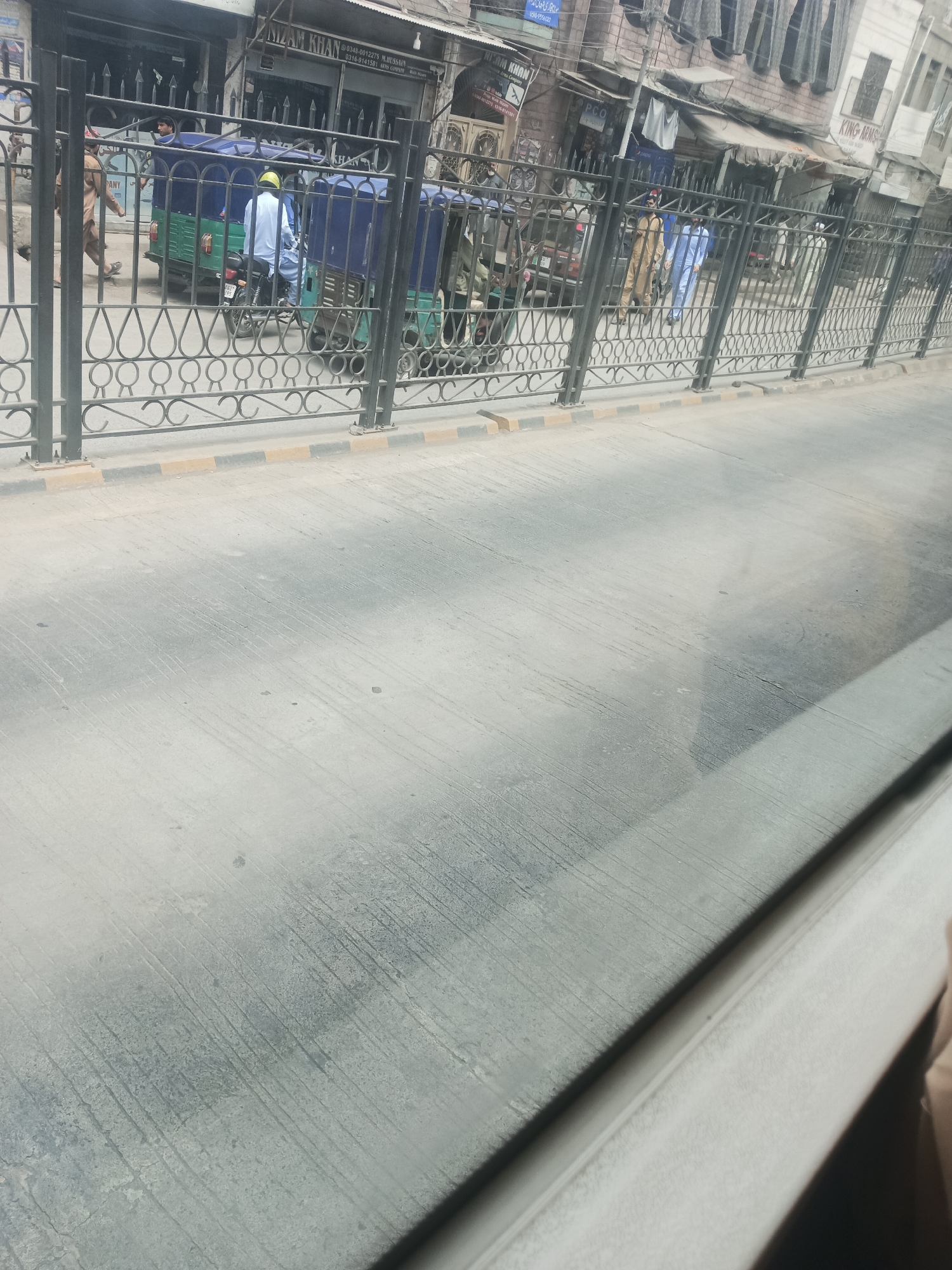 The image depicts a street scene in Alam Khan Kalay, Pakistan, viewed through a window or barrier. In the background, there are buildings with shopfronts and awnings. A black metal fence with decorative scrollwork runs across the middle ground of the image. Behind the fence, a light green and blue auto-rickshaw is parked or moving slowly. A person wearing a yellow helmet and white clothing is seated on the auto-rickshaw, likely driving it. To the left of the auto-rickshaw, a person in a light brown outfit is walking. Further to the right, behind the fence, two individuals in light blue and beige traditional clothing are visible. The foreground consists of a paved road surface with visible tire marks and debris. The overall lighting suggests daytime.