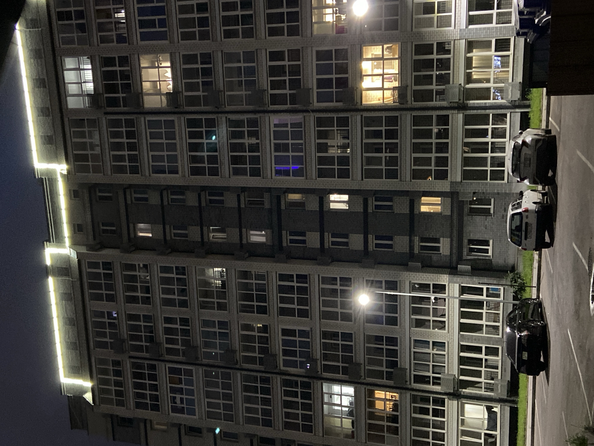 The image depicts a night-time view of a multi-story apartment building with several illuminated windows. The building appears to be constructed with light-colored bricks and features numerous balconies and large windows. Decorative yellow lights are installed along the top edge of the building. In front of the building, a parking lot is visible with three cars parked: a dark-colored coupe on the left, a white hatchback in the center, and a silver hatchback on the right. A street lamp is also present, casting light on the scene. The surrounding area is dark, suggesting it is nighttime. The scene is consistent with an urban residential environment. The location is identified as Blagoveshchensk, Russia.