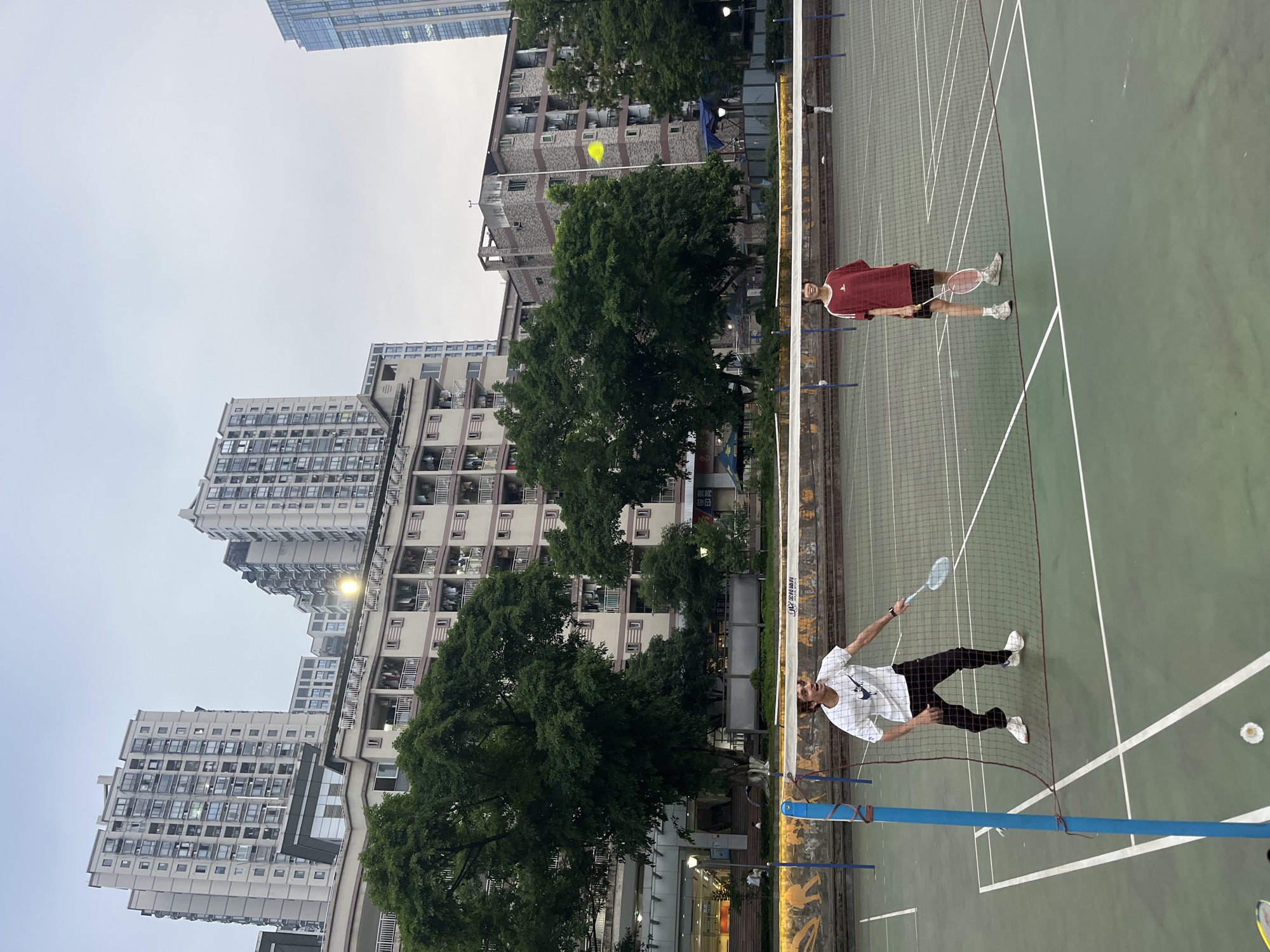 In Yuzhong District, China, two young adults are playing badminton on an outdoor court. A male, wearing a white Nike t-shirt and black pants, is mid-action, with his mouth open as if speaking or shouting. He holds a badminton racket. Facing him across the net is a female in a maroon t-shirt and black shorts, also holding a racket. The badminton court is green with white markings. In the background, several multi-story apartment buildings and trees are visible under an overcast sky. A yellow shuttlecock is in mid-air between the players, indicating an active game.