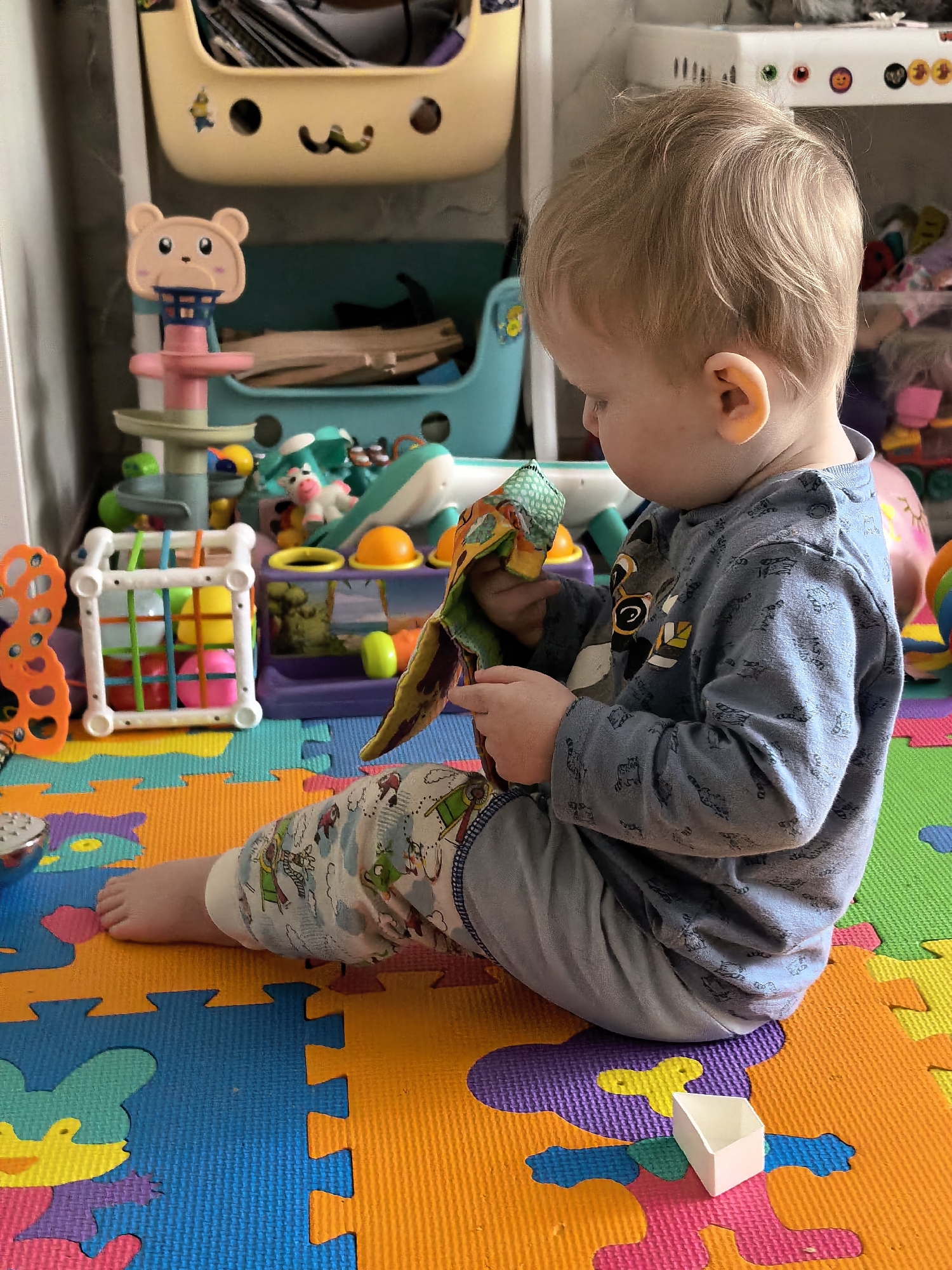 A young child with light brown hair sits on a brightly colored interlocking foam play mat, engaged in examining a vibrant fabric toy held in their hands. The child is wearing a grey-blue long-sleeved shirt with dark patterns and light-colored pants featuring cartoon rockets and stars. They are barefoot, with one foot resting on the orange and blue sections of the mat.
The scene is an indoor play area, likely a child's room, filled with an assortment of toys. In the background, to the left, a white cage-like toy holds colorful balls, next to an orange and white patterned toy. Further back, a bear-faced basketball hoop toy stands near a light blue storage bin filled with wooden blocks. Above this, a beige storage bin adorned with a cartoon face and a sticker contains various items. To the right, a purple and yellow activity toy with a landscape image is visible, along with a light blue whale-shaped toy. A small white plastic block rests on the play mat near the child's right leg.
The bright, even lighting suggests it is daytime. The child appears calm and focused on their play. This domestic setting, characterized by the play mat and numerous toys, is located in Sterlitamak, Russia. No visible text is clearly discernible within the image.