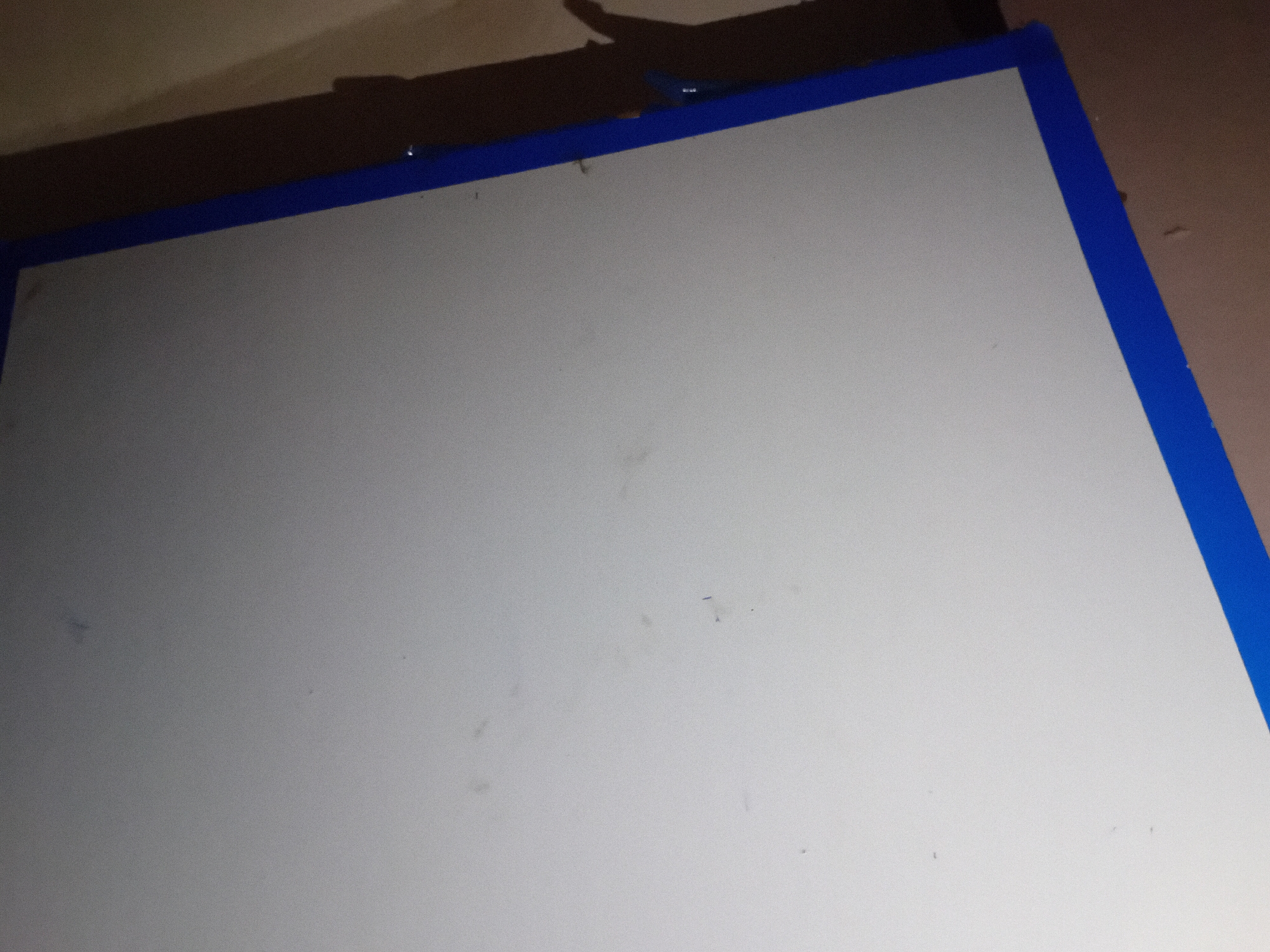 The image displays a close-up view of a large, white, flat surface that appears to be a whiteboard or a similar material. The surface is bordered by a bright blue strip along the bottom and right edges, suggesting it might be a framed board or has been taped off. There are a few faint, dark specks and smudges on the white surface, possibly dust or faint markings. The background to the right of the white surface is a wall with a textured, light brown or beige color. A shadow is cast on this wall, indicating a light source from the left, out of frame. There is a visible shadow of what appears to be a hand with outstretched fingers. The overall lighting is dim, with some areas of the white surface appearing brighter than others due to the light source. There is no discernible location in a city; it appears to be an indoor setting.