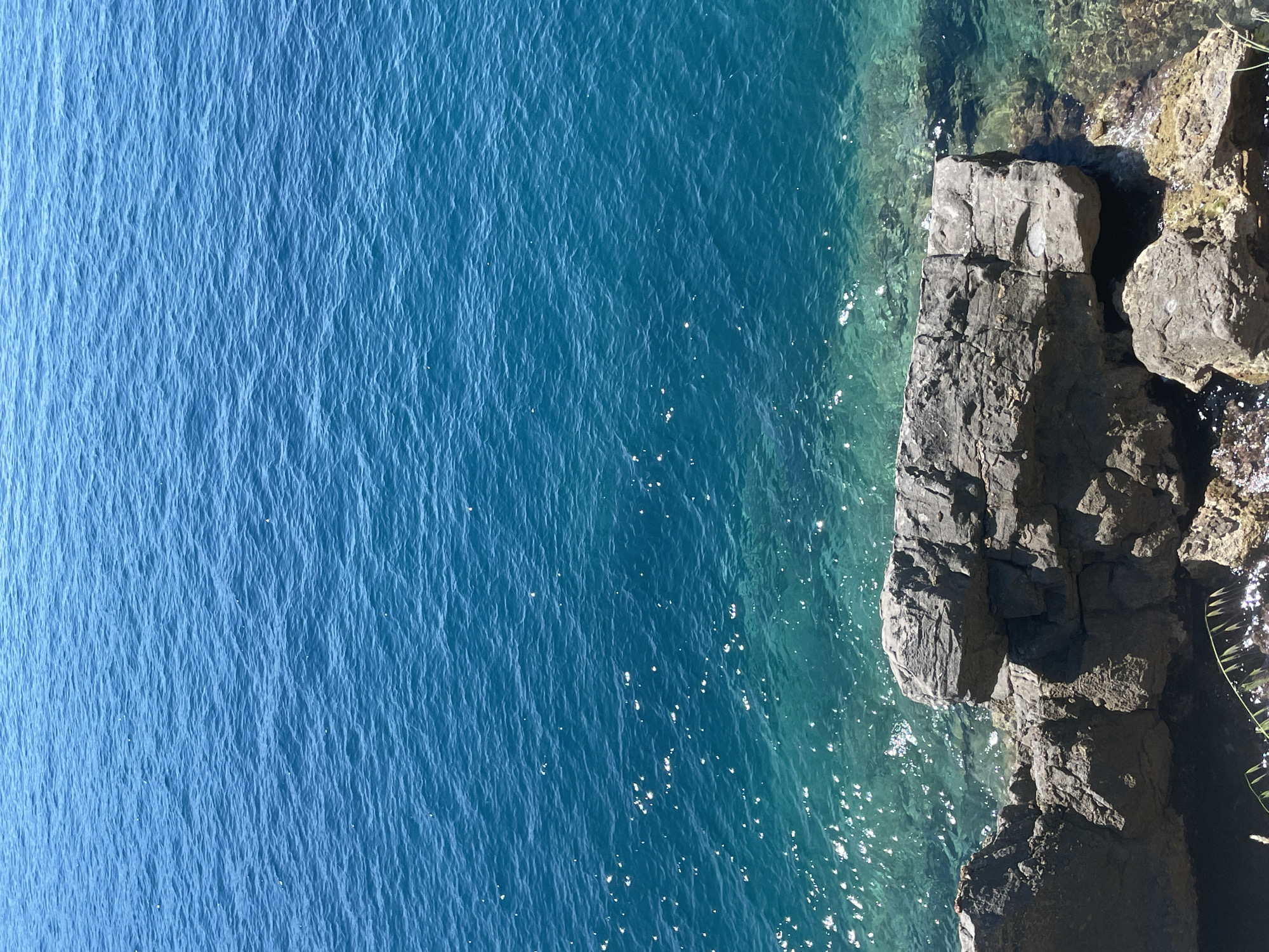 An aerial view shows a stretch of the Adriatic Sea meeting a rocky coastline. The water ranges in color from light blue to turquoise with sunlight reflecting off the surface. The rocky terrain, likely part of a cliff or outcropping, consists of gray and brown rocks. The location is likely near Boreti, Montenegro, given the presence of Adriatic Sea and rocky terrain. No people or subjects are visible in the image.