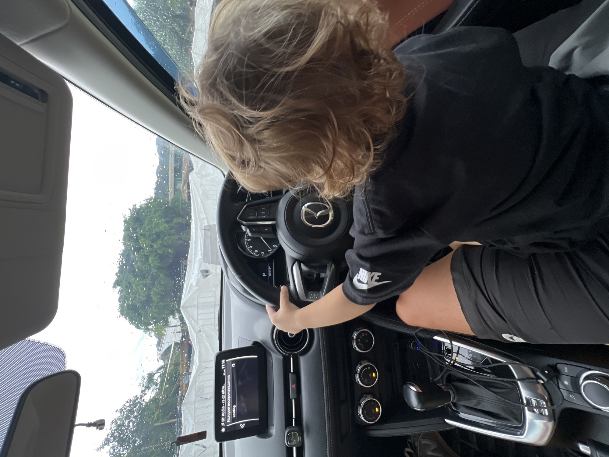 A child with blond hair is sitting in the driver's seat of a car, holding the steering wheel. The car's dashboard and controls are visible. Outside the window, there are trees and structures covered in white tarps. The car's infotainment system is displaying "BT Audio - rs gt ultra... История воспроизведения Loading". The dashboard indicates a gear selection of 'P' and displays "20/13". The child is wearing a black shirt with a white Nike logo and black shorts.