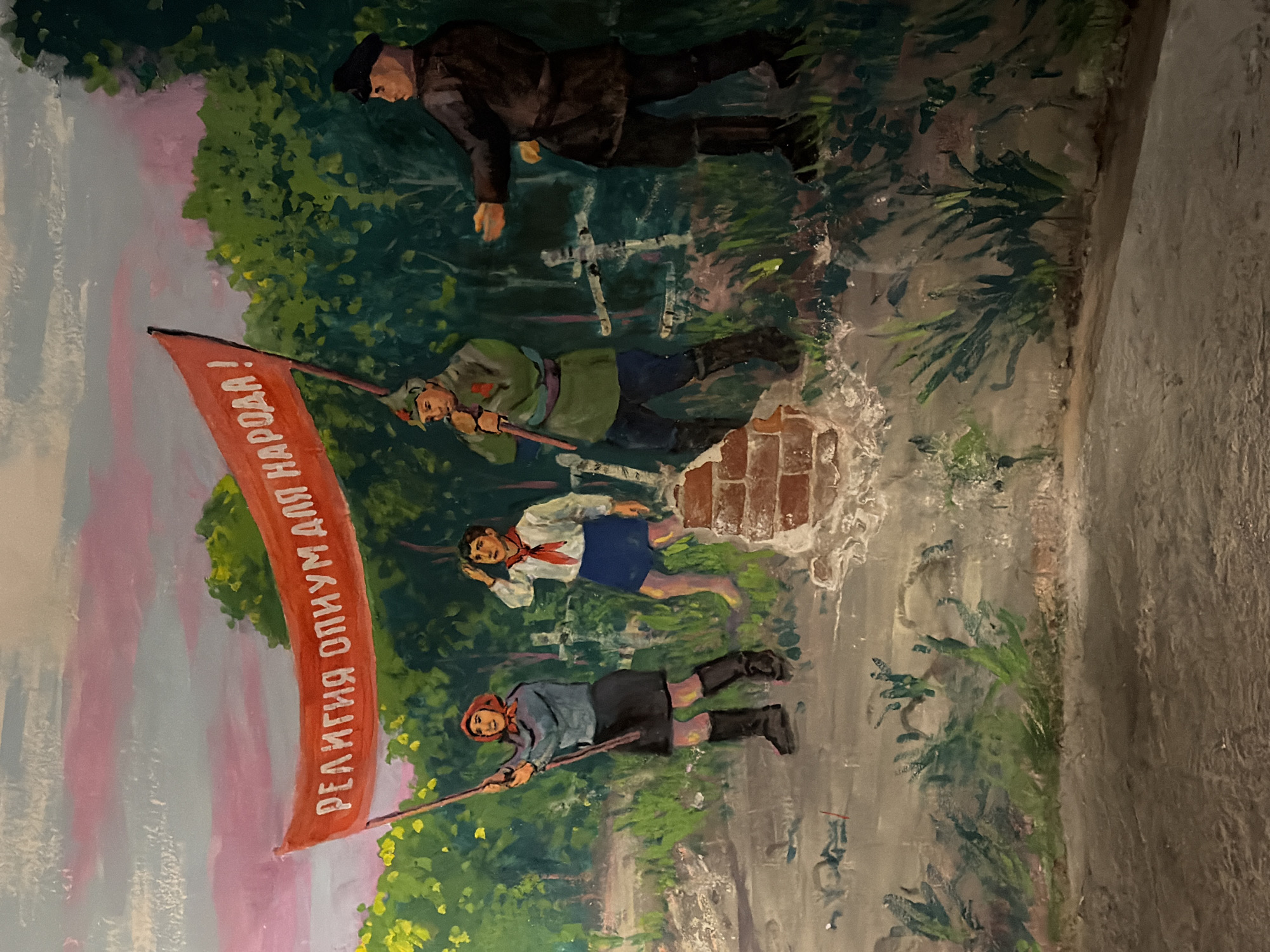 The image shows a section of a mural located in Saint Petersburg, Russia. The mural depicts figures in a socialist realist style. At the top, a man in a dark coat and cap is depicted. Below him, a man in military-style clothing carries a red flag with Cyrillic text that reads "Religion is opium for the people!". Further down, there's a child in a school uniform with a red scarf, likely a Young Pioneer, and another figure holding the flag. The background consists of trees and a sky painted with pink and white hues. The mural appears to be painted on a brick wall that is damaged.