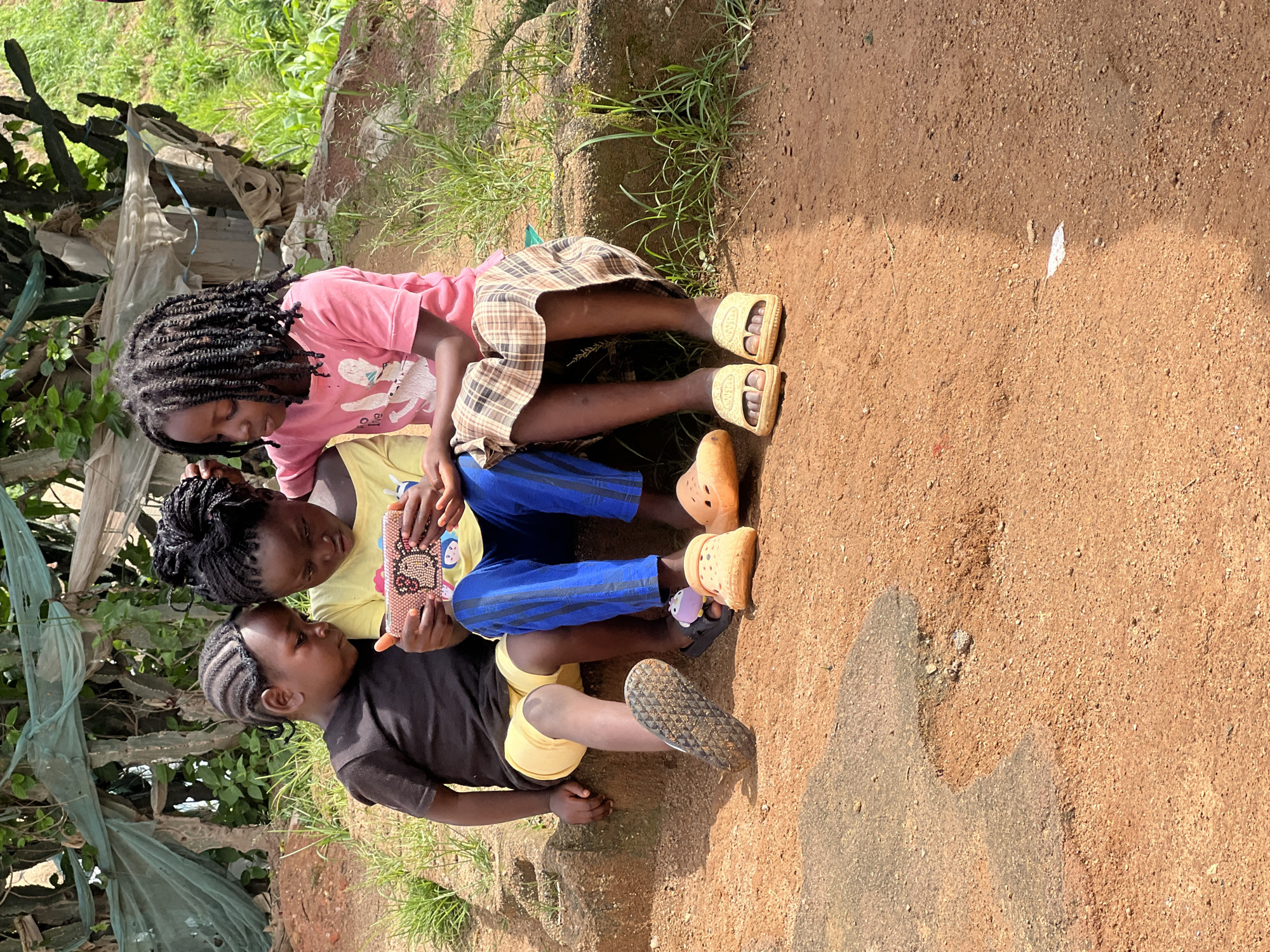 Three young Black children are sitting on a reddish-brown ledge outdoors, looking at a smartphone held by the child in the middle. The child on the left, wearing a black t-shirt and yellow shorts, has braided hair. The child in the middle, in a yellow t-shirt and blue pants, is holding the phone. The child on the right, wearing a pink t-shirt and a plaid skirt, has long, dark braided hair. All three children are wearing sandals. The background shows greenery and what appears to be a makeshift shelter. The ground is a mix of reddish-brown soil and some patches of grey concrete. The overall impression is a casual outdoor scene.