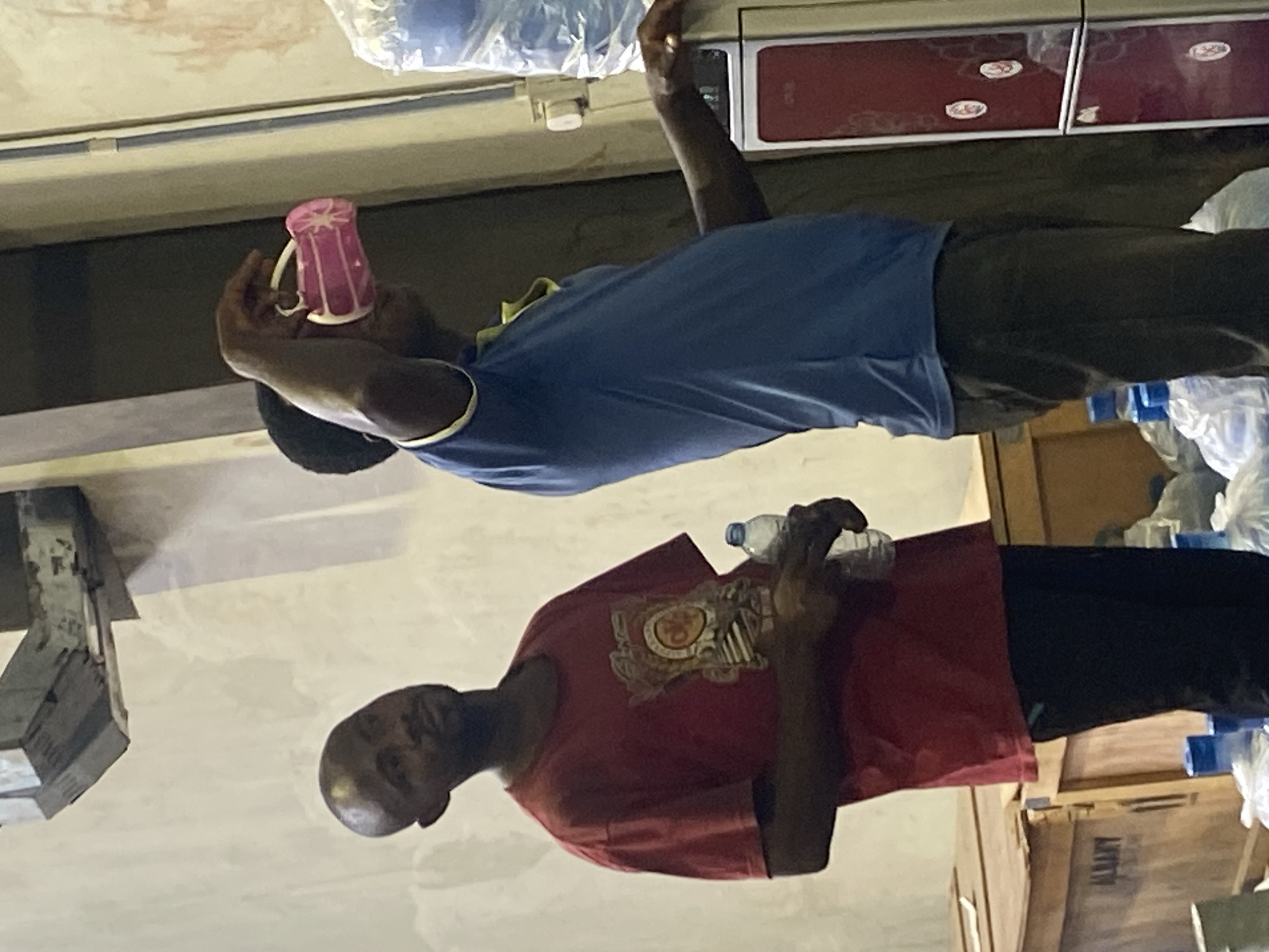 Two African males are captured in the image. One man, wearing a blue shirt and black trousers, holds a pink mug to his mouth, seemingly drinking. The other individual is dressed in a red shirt and black pants, clutching a plastic water bottle. The backdrop features water dispensers and stacked bottles of water, indicating a commercial environment. The presence of wooden crates labeled "ALMATY" suggests a distribution or storage facility. There is no location in Nigeria named Paipe. Further detail is not possible based on the image.
