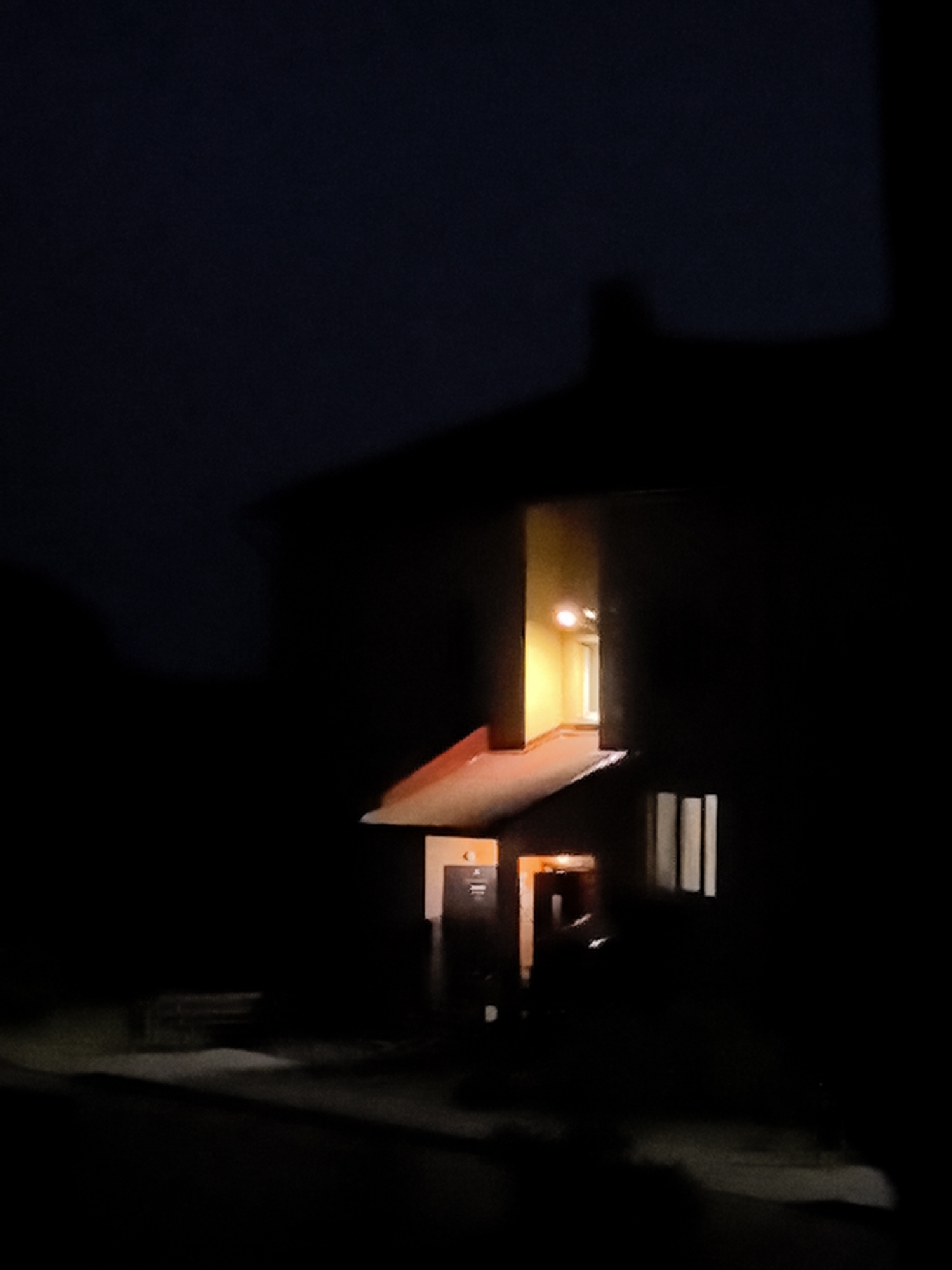 This media file is a low-light photograph of the exterior of a building at night. The building is silhouetted against a dark sky, with a warm light emanating from a window in an upper section of the structure. Below this, a covered entranceway is illuminated, casting a reddish-orange glow. The entrance features a dark door and to its right, a vertical sign with what appears to be a symbol and some text, though it is largely unreadable due to the low light and resolution.
To the left of the entrance, the outline of a bench is visible in the darkness, suggesting an outdoor seating area. The ground in front of the building is indistinct but appears to be a pathway or paved area, with shadows cast across it. Vertical window panes are visible on the right side of the building, also dimly lit from within. The overall impression is one of quiet isolation, with the lit interior offering a sense of occupancy against the dark, perhaps desolate, surroundings. No people are visible in the photograph. The time of day is clearly night, and no weather conditions are discernible.