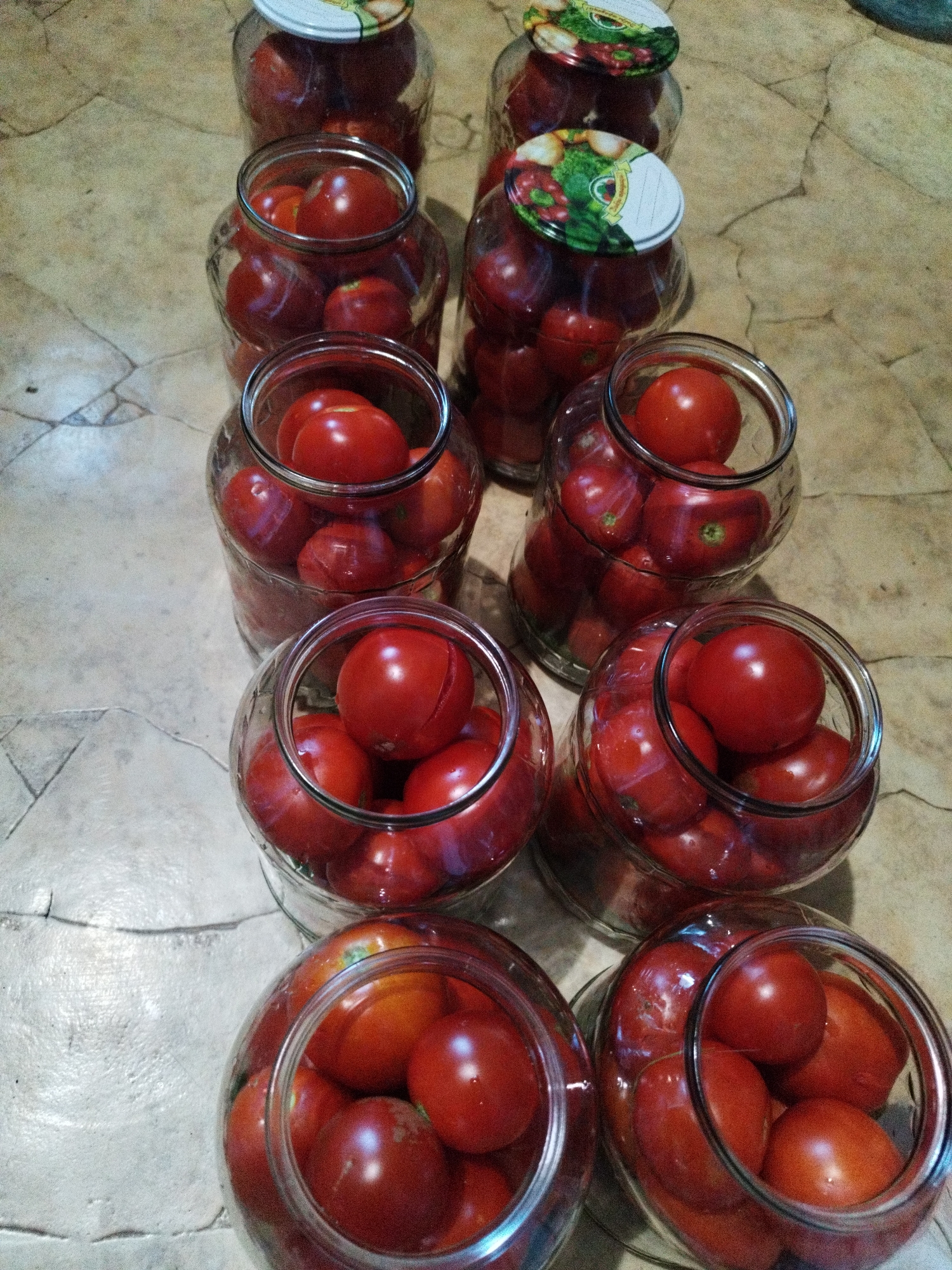 This is a close-up, high-angle photograph of several glass jars filled with ripe red tomatoes, arranged in rows on a textured, light-colored surface, likely a countertop or table. The jars vary in fullness, with some packed tightly and others less so. The lids of two jars are partially visible, one with a decorative label featuring an illustration of vegetables. The lighting in the scene is somewhat dim, casting soft shadows and highlighting the glossy surface of the tomatoes and glass. There are no people or animals present in the image. The setting appears to be a domestic kitchen. The location is specified as Lozova, Ukraine.