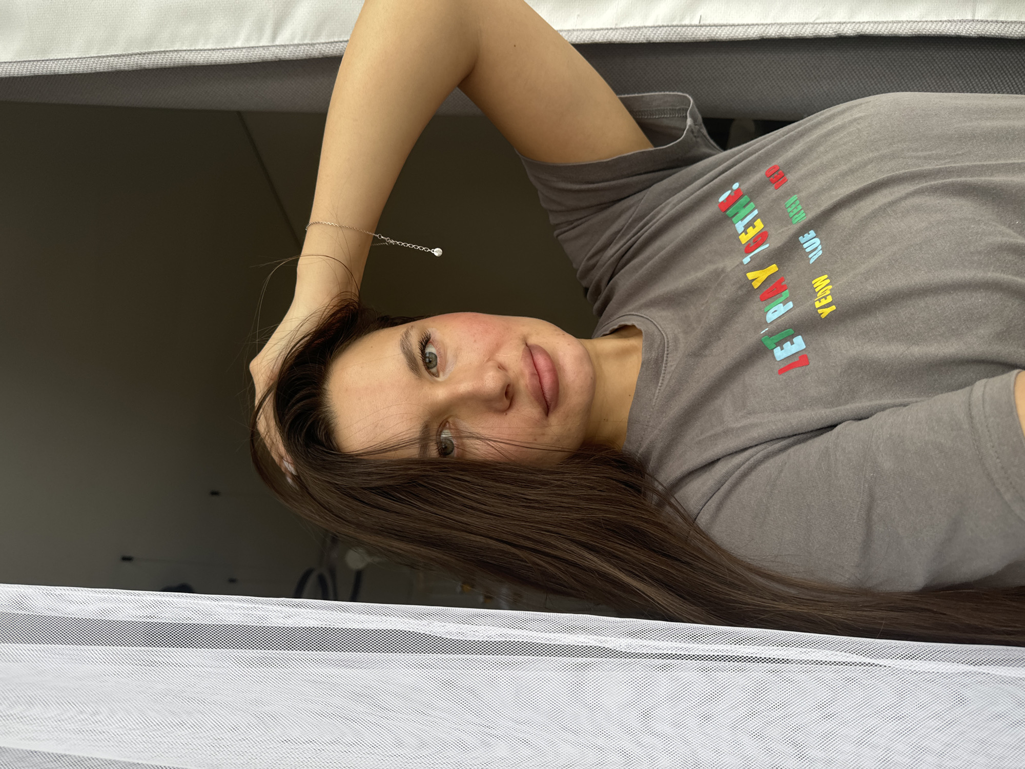 A young woman is depicted lying down in an indoor setting, looking directly at the camera from a low angle. She is positioned on her side with her left arm raised, her hand resting on her long, dark brown hair. Her face is turned slightly towards the viewer, revealing light-colored eyes and a neutral expression with a hint of a smile. Her cheeks show a natural flush.
She is wearing a plain grey t-shirt that features colorful text on the front. A delicate silver necklace with a small, clear pendant hangs from her neck, visible on her raised arm. The text on her shirt reads "LET'S PLAY TOGETHER!" in large, multi-colored letters. Below this, smaller words "YELLOW", "BLUE", "RED", and "GREEN" are visible, each printed in its respective color, though "RED" and "GREEN" are partially obscured.
The woman appears to be lying on a light-colored, upholstered surface, possibly a bed or couch, visible at the top and bottom of the frame. The background is dark and indistinct, suggesting a room interior, while a white, finely meshed fabric, likely a curtain or net, occupies the lower foreground. The scene takes place in Odintsovo, Russia.