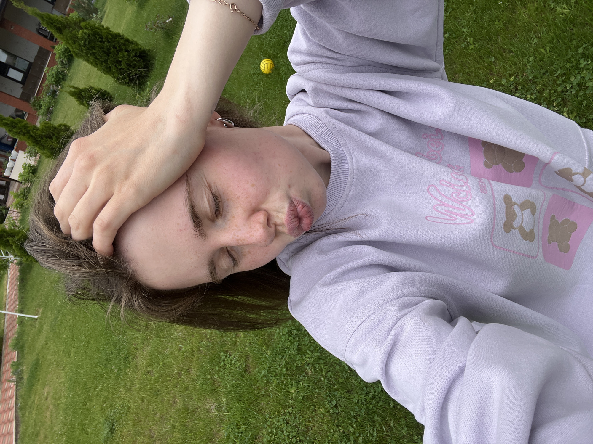 A person is on grass outdoors, wearing a light purple sweatshirt with a design of bears. The person has brown hair and is holding their hand on their forehead. They have freckles and are making a kissing face. In the background, there are green trees and grass and a building. A yellow ball is present on the grass. The location is in Chaikovsky, Russia.