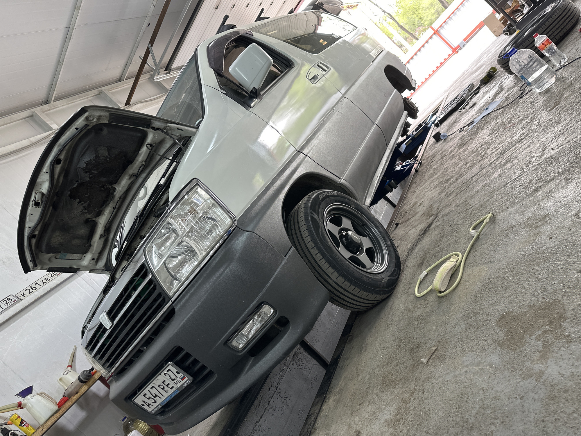 The image shows a silver van undergoing maintenance in what appears to be a garage setting. The hood is open, revealing the engine compartment. A license plate is visible on the front of the van. There are multiple bottles of fluids on a shelf in the background. The van is jacked up, with the front driver-side wheel installed. The wheel on the passenger side has been removed and a tire and some tools can be seen on the ground beside a water bottle. The photograph was taken in Blagoveshchensk, Russia, based on the license plate registration code.