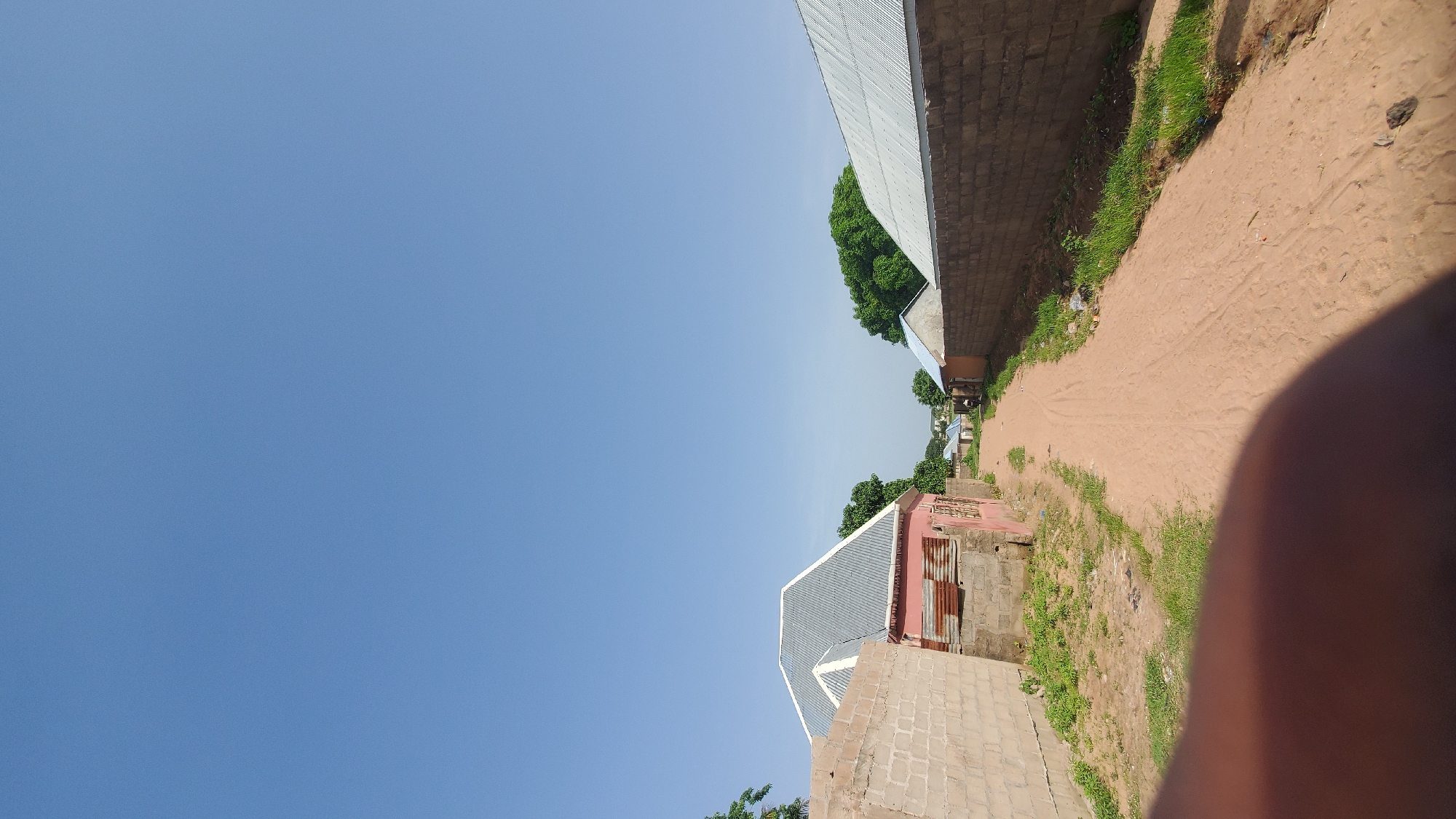 This is a vertical, eye-level, outdoor photograph taken on a sunny day in Jalingo, Nigeria. The scene depicts a dusty, unpaved road that curves to the right, leading into the distance. On the left side of the road, there is a low, light-pink building with a grey corrugated metal roof, bordered by a wall constructed of concrete blocks. Adjacent to this building, on the far left, is a taller, beige concrete block wall. On the right side of the road, there is a long, grey concrete block wall, and beyond that, a building with a light-blue metal roof. Lush green trees are visible in the background. A person's thumb and forefinger are partially visible in the lower foreground, obscuring the bottom portion of the image.